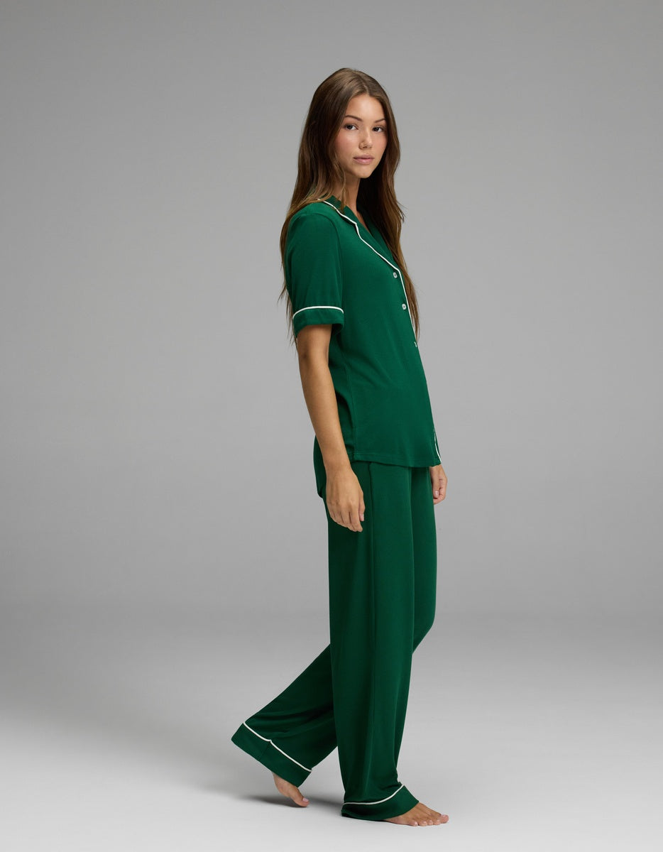A woman stands barefoot against a plain gray background, looking at the camera with a relaxed expression, wearing Cozy Earth’s Women’s Bamboo Rib-Knit Classic Short Sleeve & Pant Pajama Set in green with white trim.