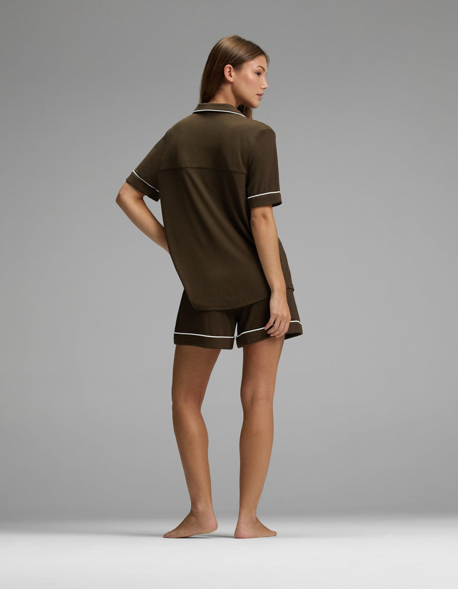 A person stands barefoot on a plain gray surface, facing away, wearing the Cozy Earth Women’s Bamboo Rib-Knit Classic Short Sleeve Pajama Set in dark brown with light trim.