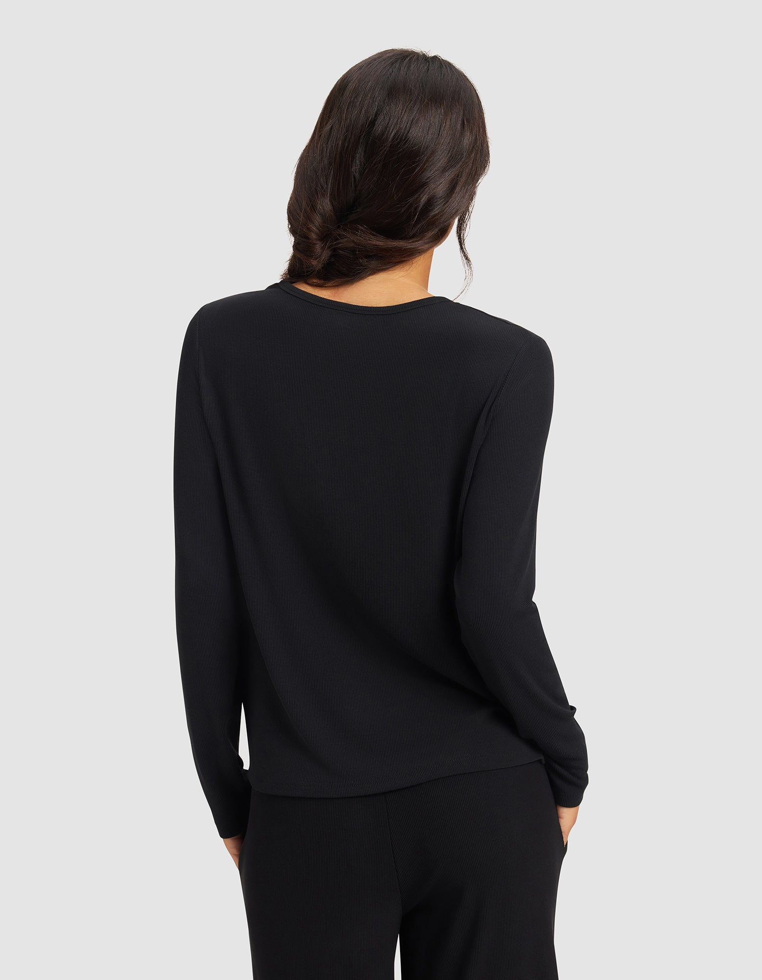 A woman with long, wavy dark hair stands with her back to the camera, wearing the Cozy Earth Women’s Bamboo Rib-Knit Long Sleeve Lounge Top in black against a plain light gray background.
