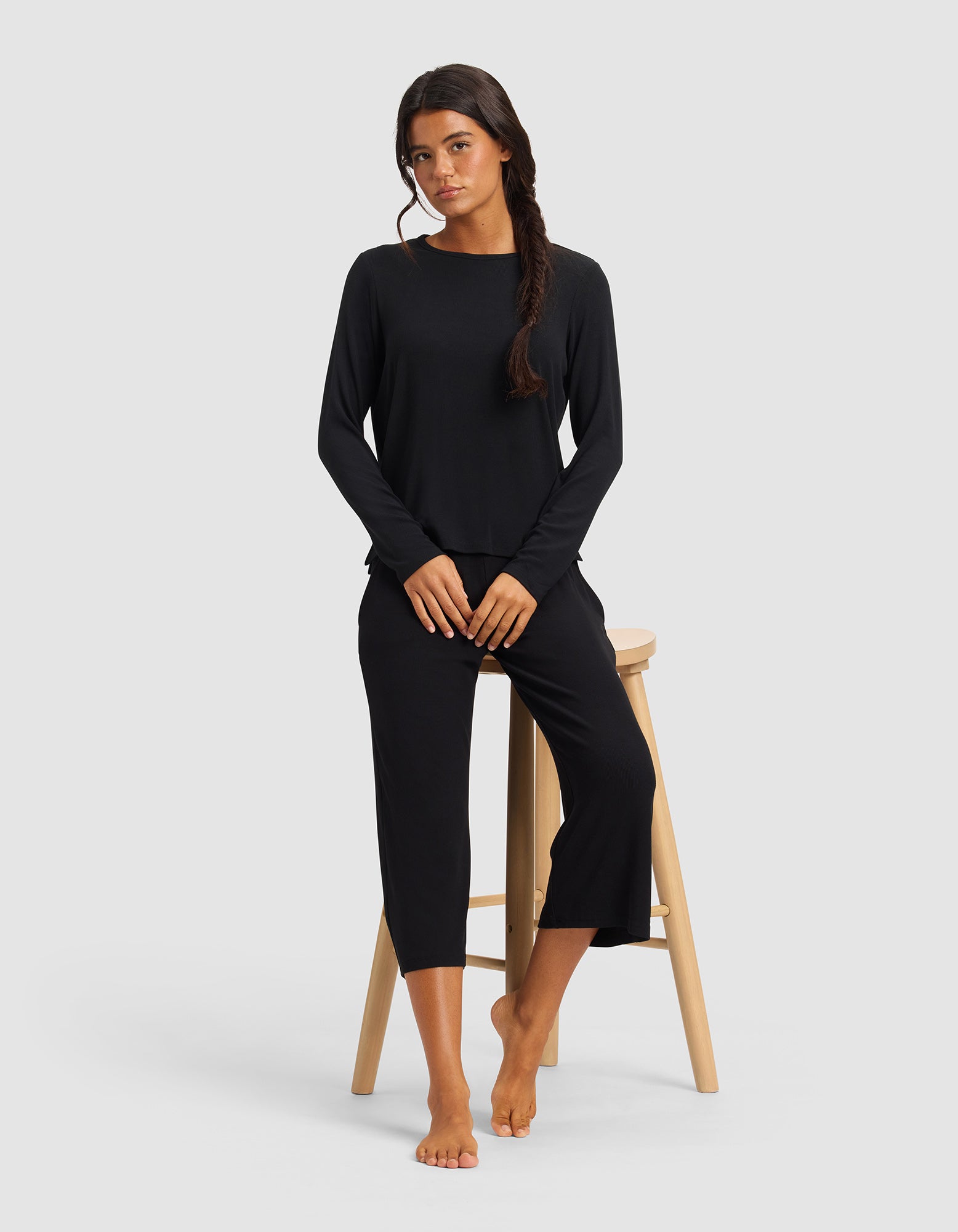 A woman with long dark hair in a loose braid wears the Cozy Earth Women’s Bamboo Rib-Knit Long Sleeve Lounge Top in black, standing barefoot beside a light wooden stool against a plain light gray background.