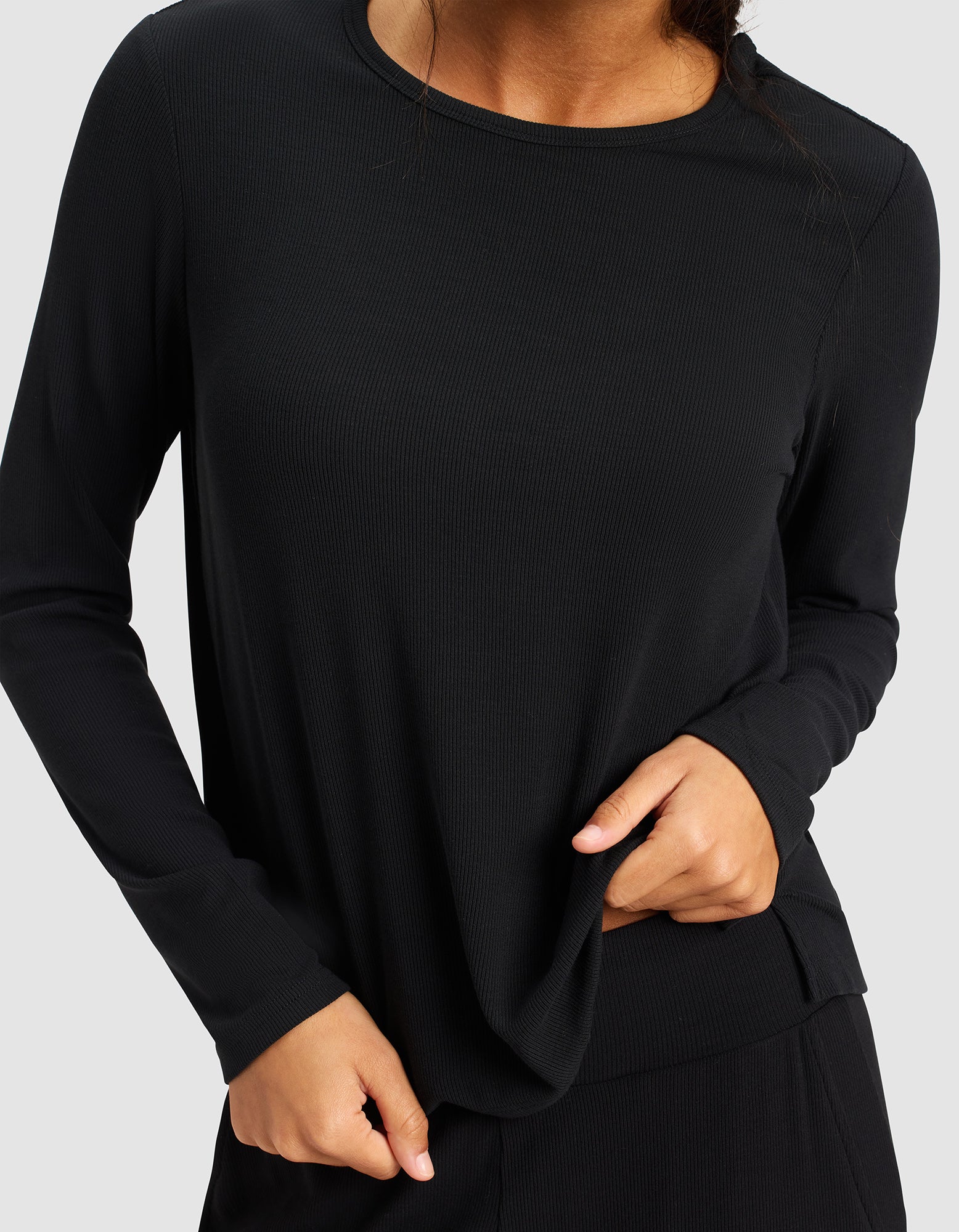 A woman models the Cozy Earth Women’s Bamboo Rib-Knit Long Sleeve Lounge Top in black, tugging slightly at the hem. Her face is partially out of frame against a plain light gray background.