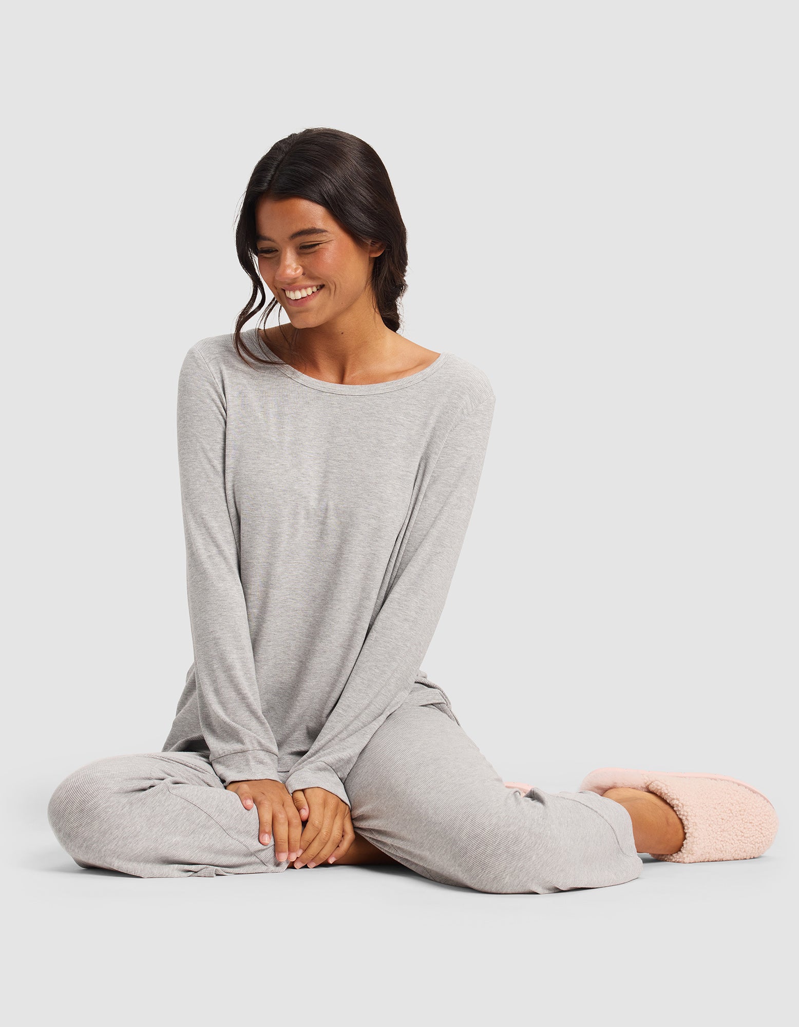 A woman with long dark hair sits cross-legged and smiles, wearing the Cozy Earth Women’s Bamboo Rib-Knit Long Sleeve Lounge Top in light gray, paired with pink fuzzy slippers against a plain light gray background.