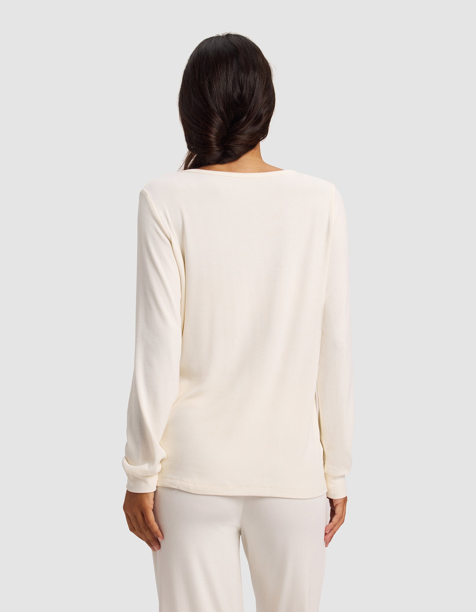 A woman with long, dark braided hair is seen from behind wearing the Cozy Earth Women’s Bamboo Rib-Knit Long Sleeve Lounge Top in off-white, paired with matching pants against a plain light gray background.
