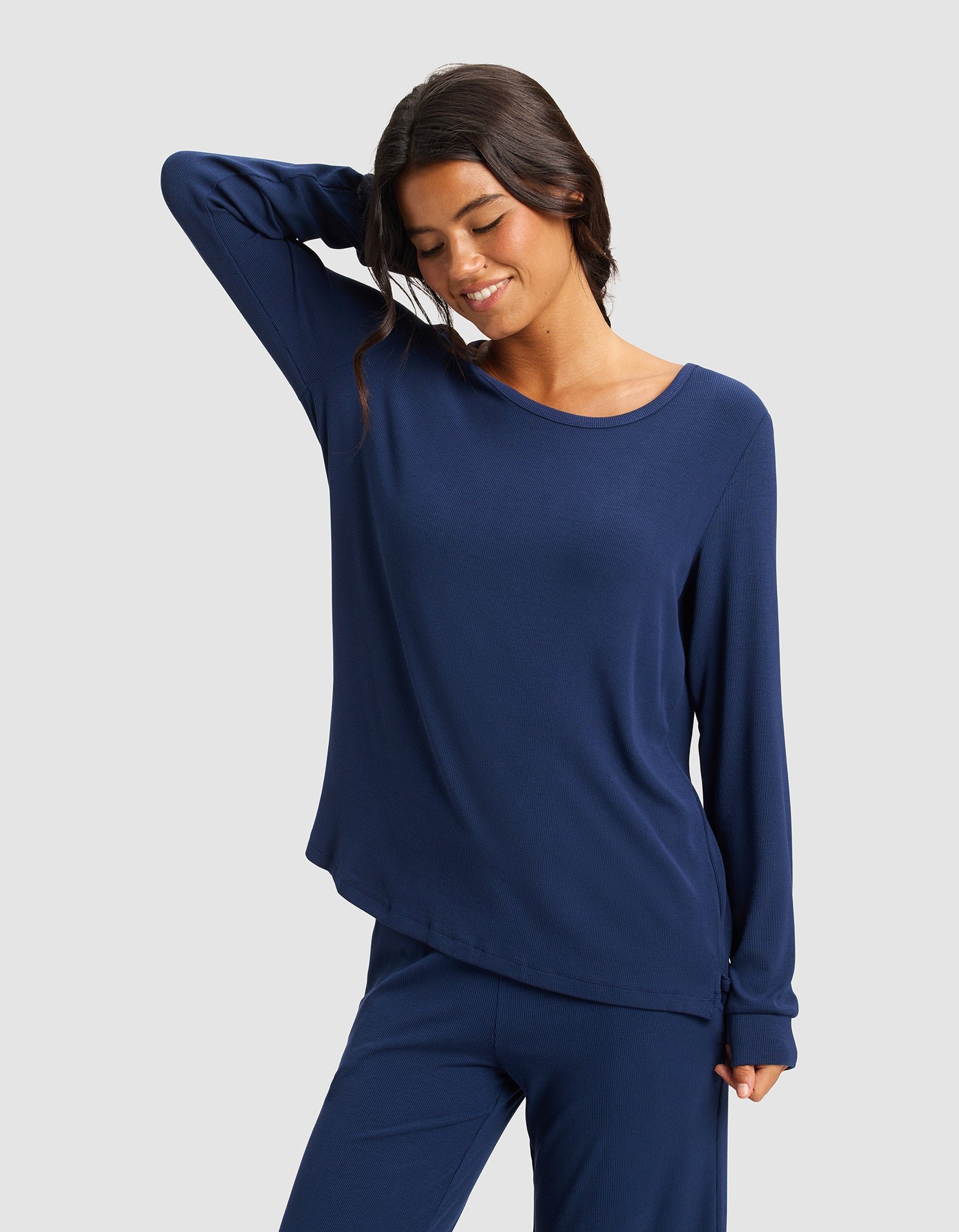 A woman wears the Cozy Earth Women’s Bamboo Rib-Knit Long Sleeve Lounge Top in navy blue, paired with matching pants, smiling and posing against a plain light gray background.|Color:Navy