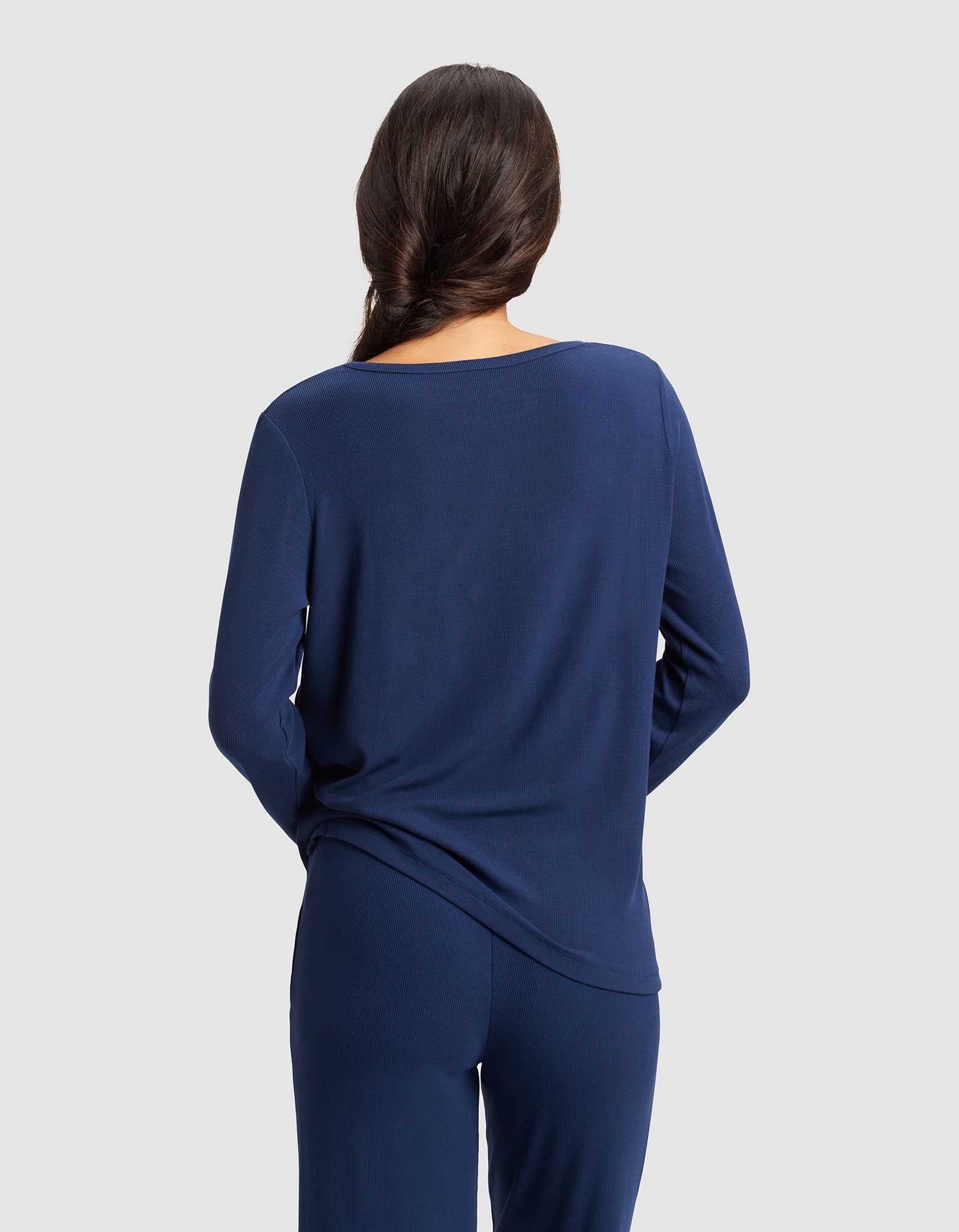 A woman with long dark hair in a loose braid stands with her back to the camera, wearing the Cozy Earth Women’s Bamboo Rib-Knit Long Sleeve Lounge Top in navy, set against a plain light gray background.