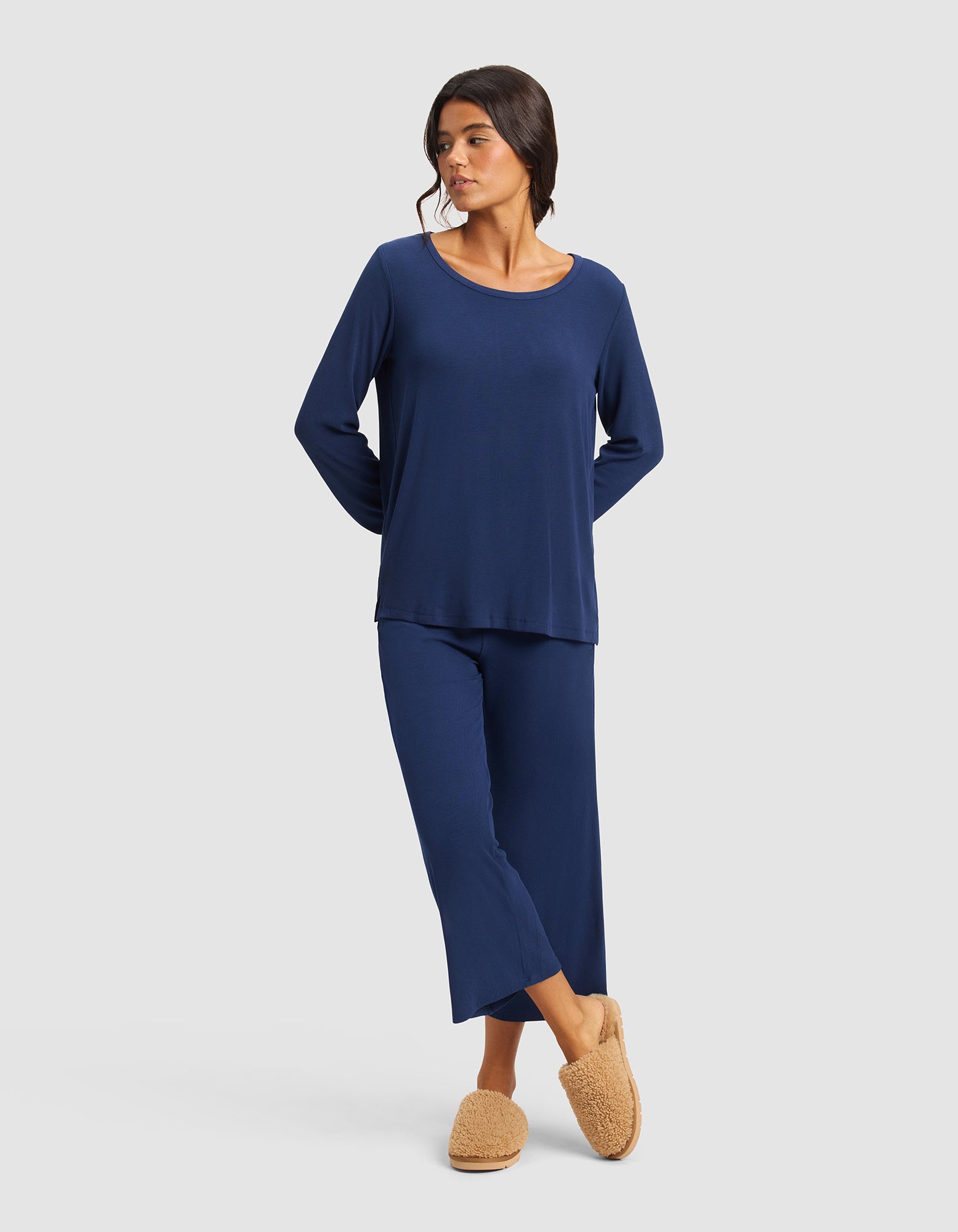 A woman wearing the Cozy Earth Women’s Bamboo Rib-Knit Long Sleeve Lounge Top in navy and tan fuzzy slippers stands with her hands behind her back, looking to the side against a plain light gray background.