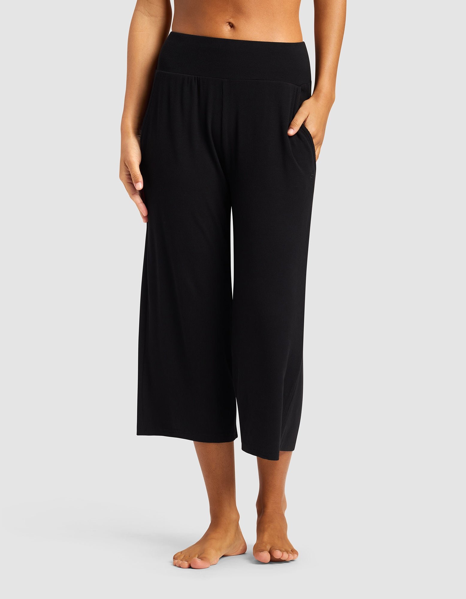 A person stands barefoot in Cozy Earth’s Women’s Bamboo Rib-Knit Lounge Capri—black, wide-leg, cropped pants with a high waistband and a pocket—against a plain light gray background.