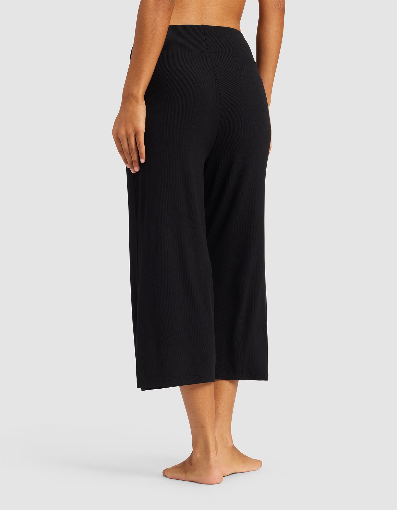 A person stands barefoot on a light background, wearing Cozy Earth’s Women’s Bamboo Rib-Knit Lounge Capri. Seen from behind, the relaxed fit and wide legs of the black cropped pants are highlighted. The upper body is mostly out of frame.