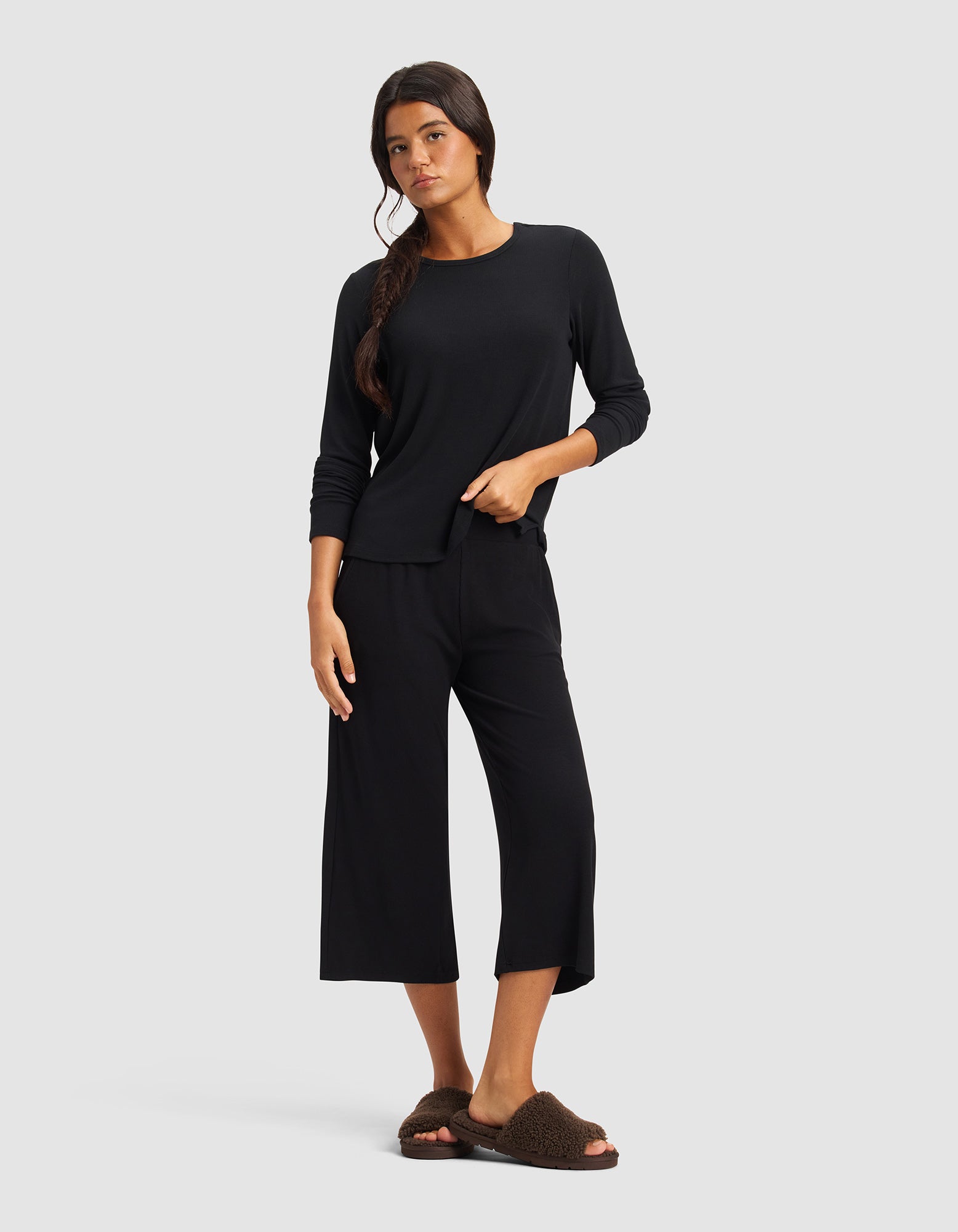 A woman with long dark hair in a loose side braid stands against a plain background wearing Cozy Earth’s Women’s Bamboo Rib-Knit Lounge Capri, a black long-sleeve top, and brown fuzzy slippers.