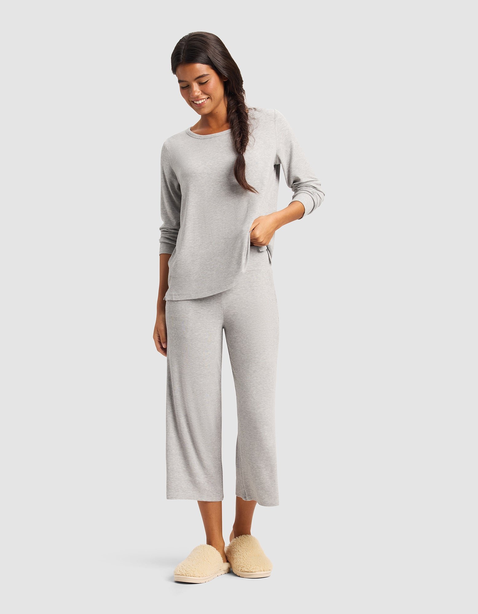 A woman with long dark hair in a loose braid smiles while wearing Cozy Earth’s Women’s Bamboo Rib-Knit Lounge Capri set in light gray and beige fuzzy slippers, standing against a plain light background.