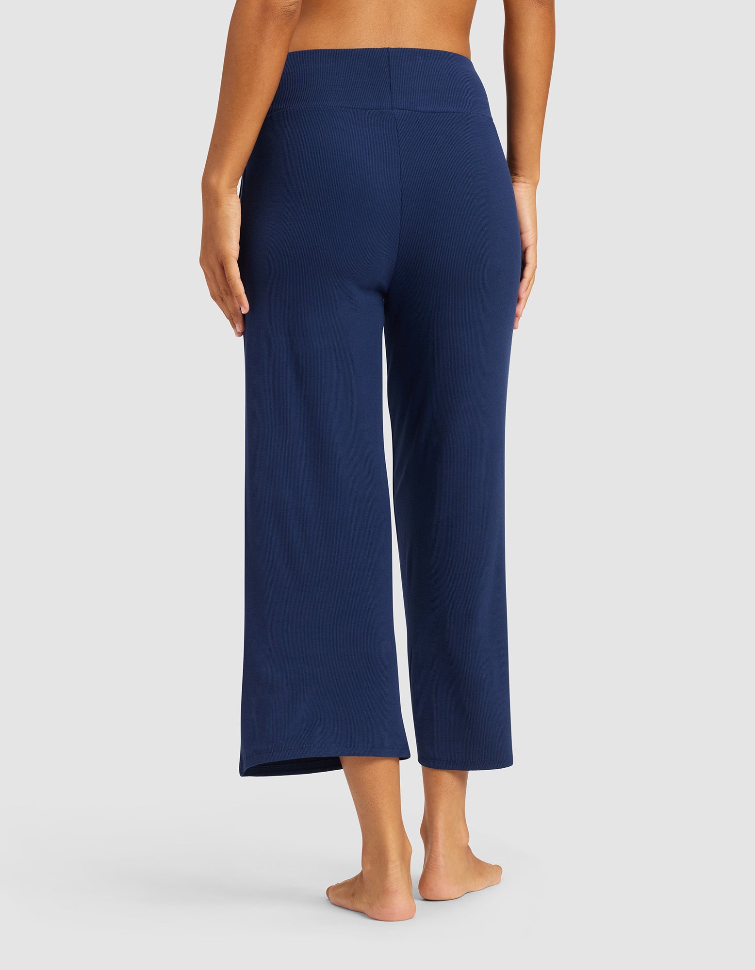 A person stands barefoot, facing away, wearing Cozy Earth Women’s Bamboo Rib-Knit Lounge Capri in navy blue. The pants are wide-leg and cropped. The background is plain light gray.