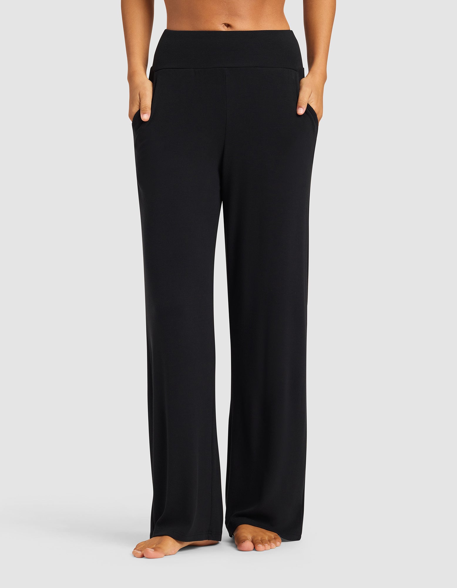 A woman stands barefoot against a light gray background, wearing Cozy Earth’s Women’s Bamboo Rib-Knit Lounge Pant in black, featuring a relaxed fit, wide waistband, pockets, and rib-knit texture for comfort.|Color:Black