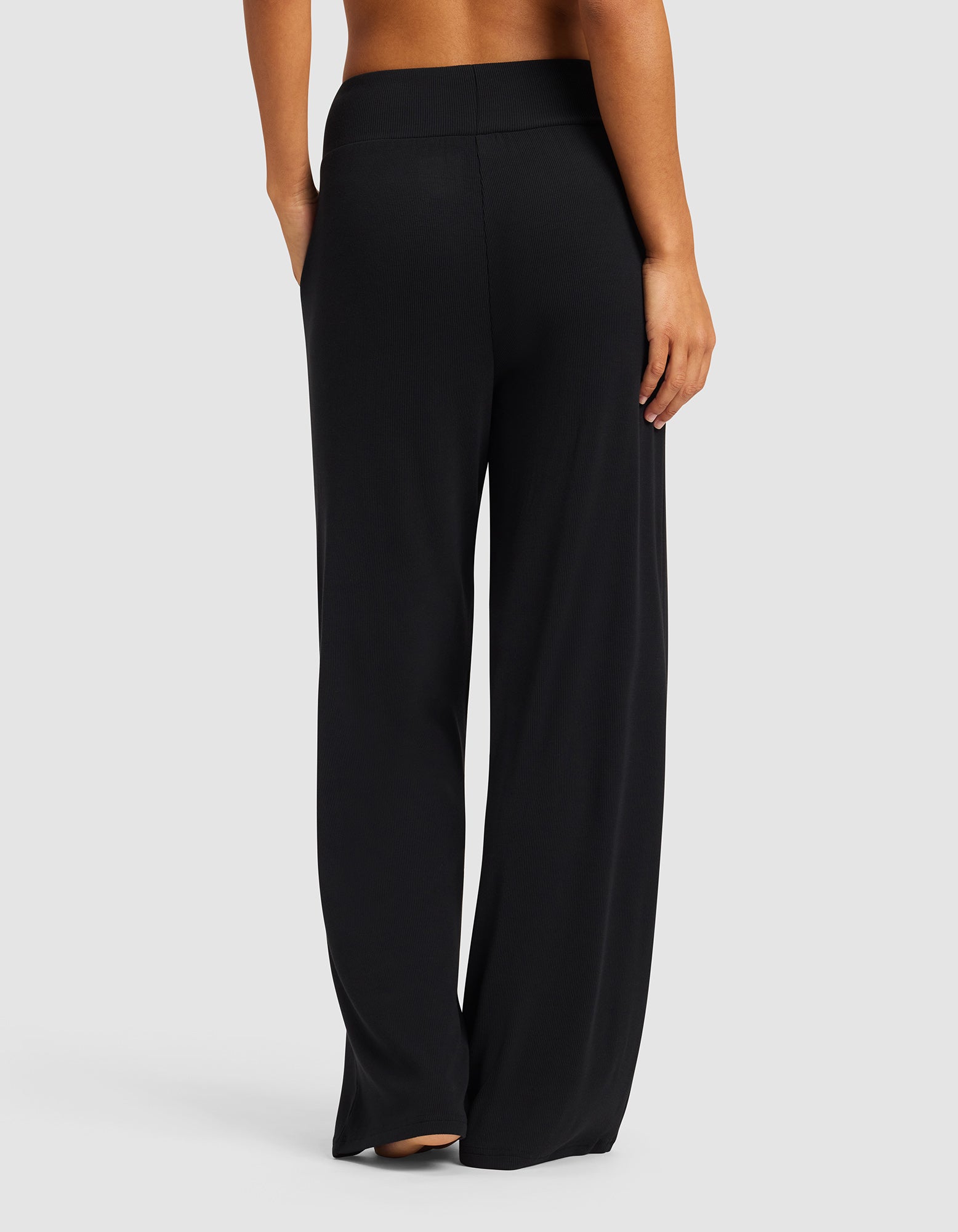 A person stands barefoot facing away, wearing Cozy Earth’s Women’s Bamboo Rib-Knit Lounge Pant with a high waistband, against a plain light gray background.