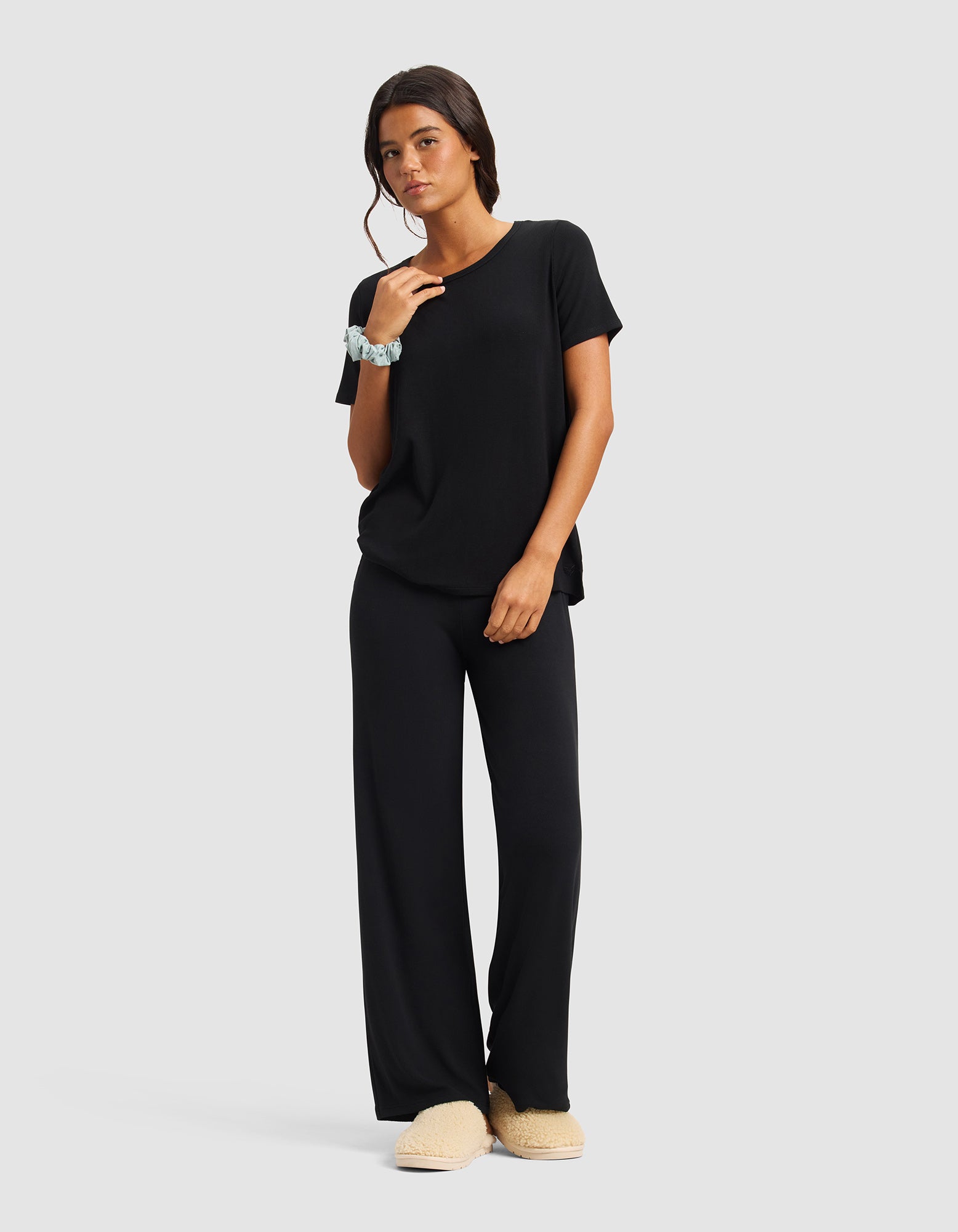 A woman with dark wavy hair stands against a plain background wearing Cozy Earth's Women’s Bamboo Rib-Knit Lounge Pant in black, a matching loose T-shirt, beige slippers, and a light blue scrunchie on her wrist.