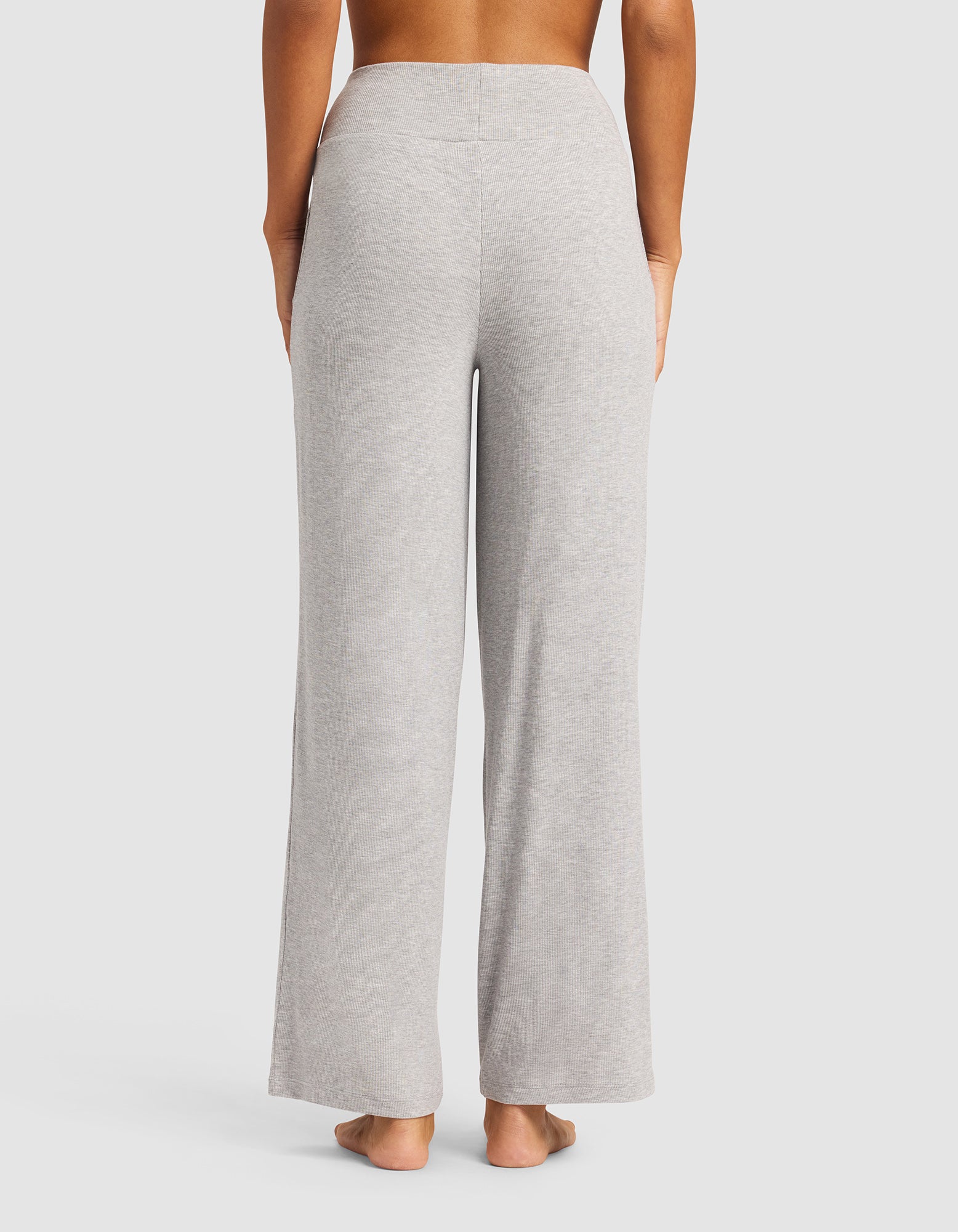 A person stands barefoot, facing away from the camera, wearing Cozy Earth Women’s Bamboo Rib-Knit Lounge Pant in light gray with a high-waisted, wide-leg fit against a plain light background.