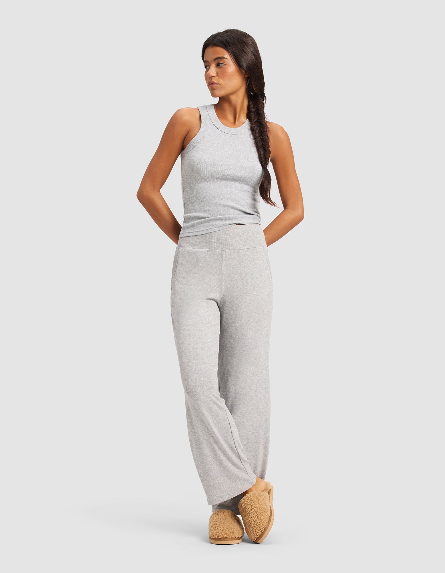 A woman with long dark hair in a braid models Cozy Earth’s Women’s Bamboo Rib-Knit Lounge Pant in light gray, paired with a matching sleeveless top and tan slippers, standing against a plain light background.
