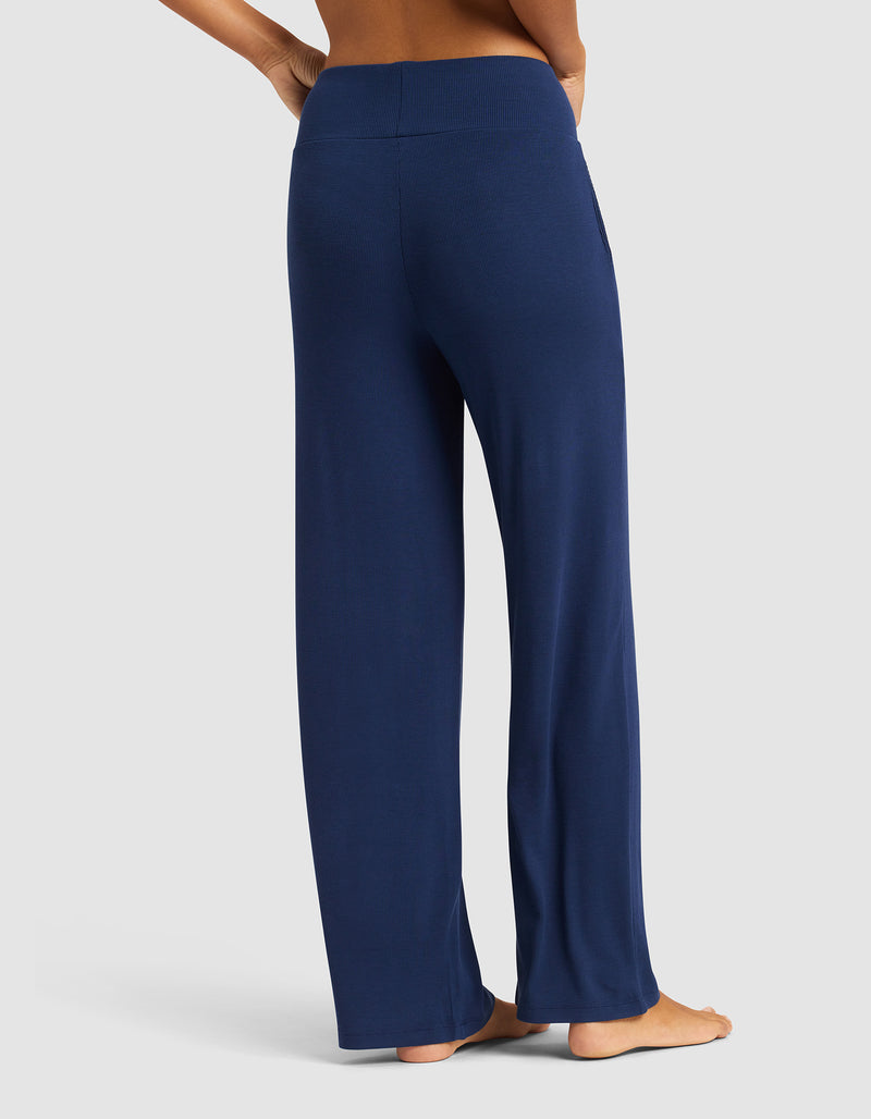 A woman models Cozy Earth’s Women’s Bamboo Rib-Knit Lounge Pant in navy, featuring a relaxed fit and high waistband, standing barefoot on a plain light gray background. The image is taken from the back, highlighting the flowy style.