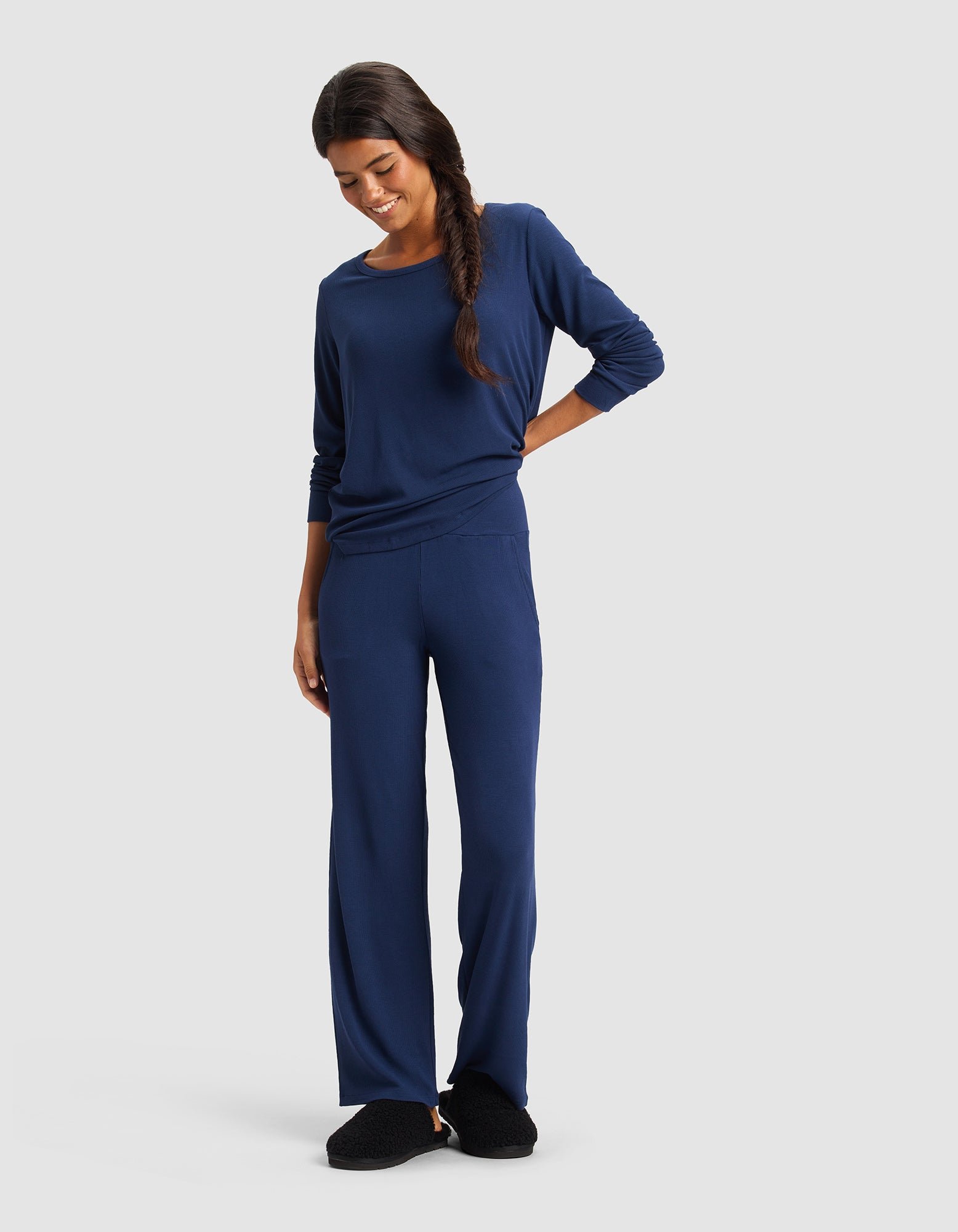 A woman with long dark hair in a side braid wears Cozy Earth Women’s Bamboo Rib-Knit Lounge Pant in navy blue with a matching top and black slippers, standing and smiling against a plain light background.|Color:Navy