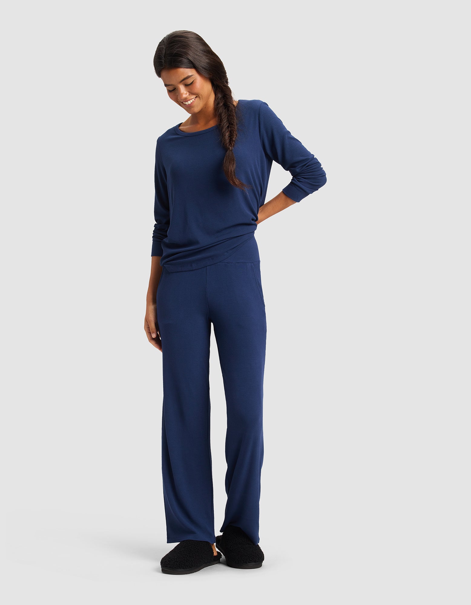 A woman with long dark hair in a side braid wears Cozy Earth Women’s Bamboo Rib-Knit Lounge Pant in navy blue with a matching top and black slippers, standing and smiling against a plain light background.|Color:Navy