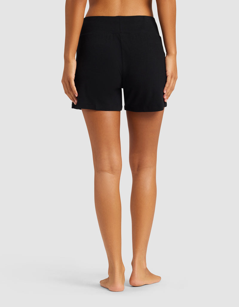 A person stands barefoot with their back to the camera, wearing Cozy Earth Women’s Bamboo Rib-Knit Lounge Shorts in black, no top, against a plain light gray background.