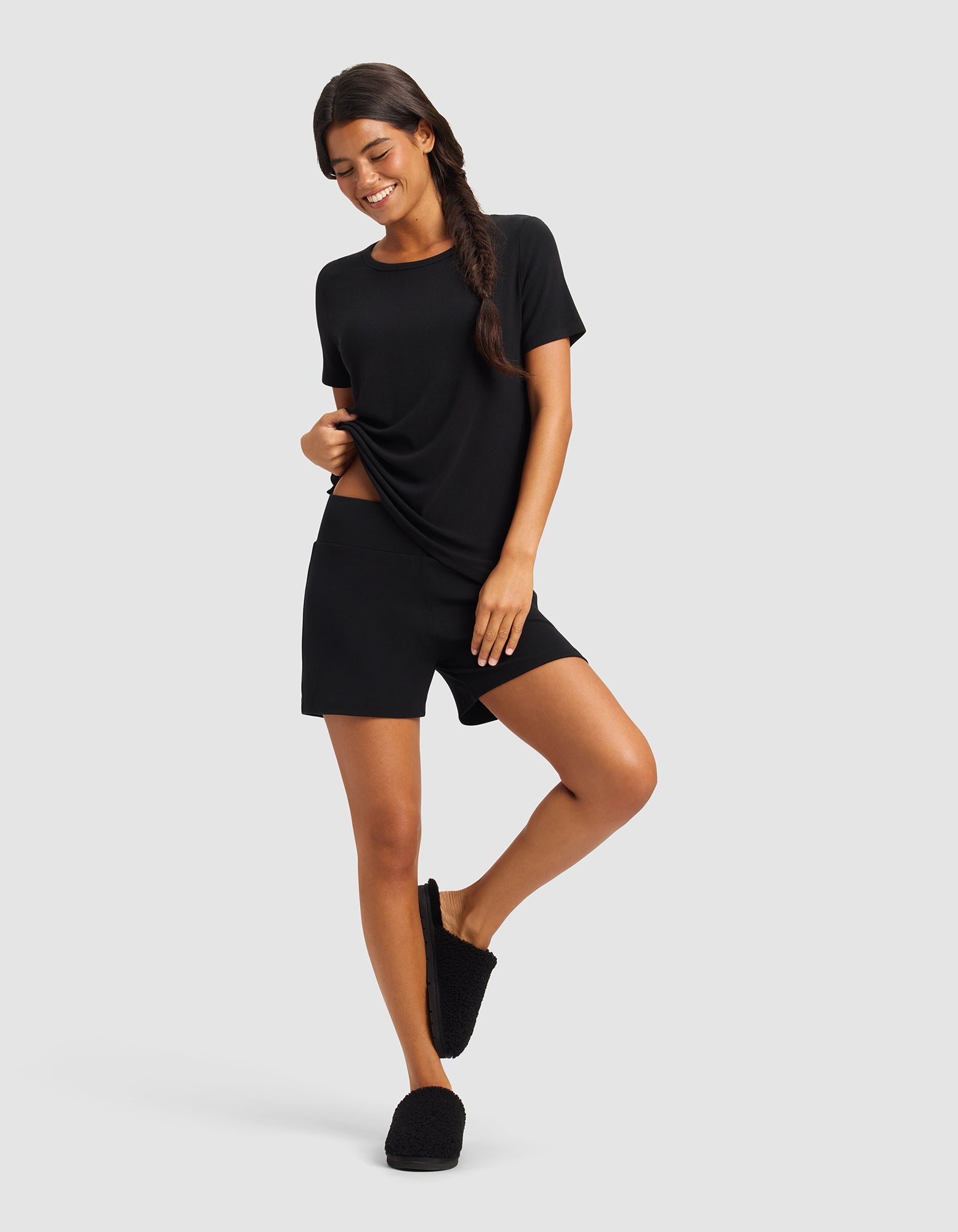 A woman poses against a light gray background wearing Cozy Earth's Women’s Bamboo Rib-Knit Lounge Short in black, along with a matching black t-shirt and slippers. She smiles, has long dark hair in a side braid, and stands with one hand on her hip.