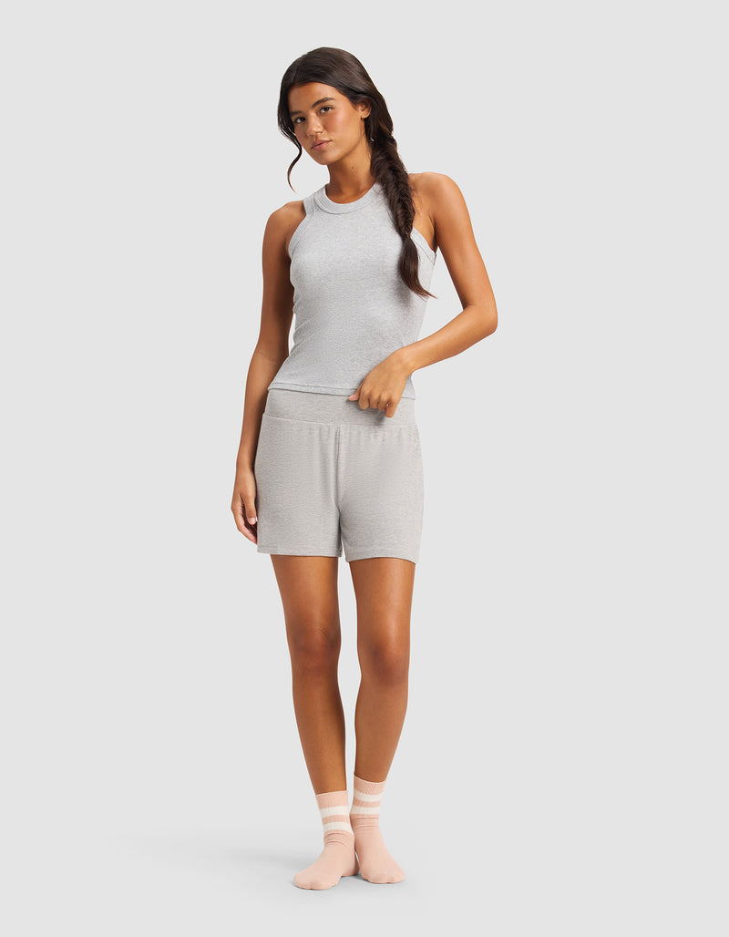A woman with long dark hair in a loose side braid poses in a relaxed stance, wearing Cozy Earth’s Women’s Bamboo Rib-Knit Lounge Short against a plain background. She pairs the shorts with a light gray tank and pale pink socks.