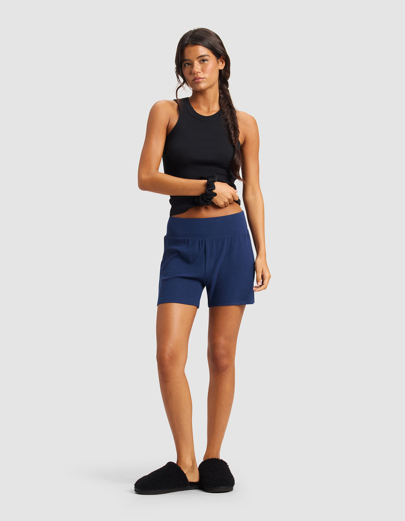 A woman with long brown hair in a side braid wears Cozy Earth’s Women’s Bamboo Rib-Knit Lounge Short in navy blue, a black sleeveless top, black slippers, and a black scrunchie on her wrist, standing against a plain light gray background.