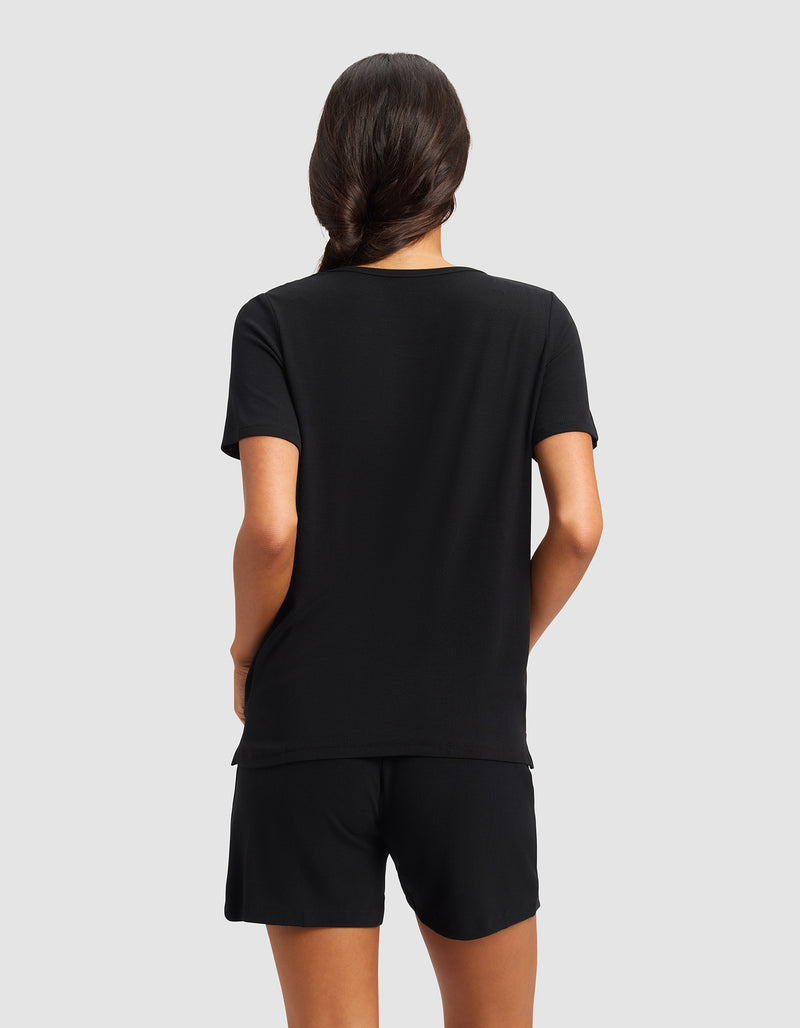 A person with long dark hair in a low braid is shown from behind, wearing the Cozy Earth Women’s Bamboo Rib-Knit Short Sleeve Lounge Top in black and black shorts against a light gray background.
