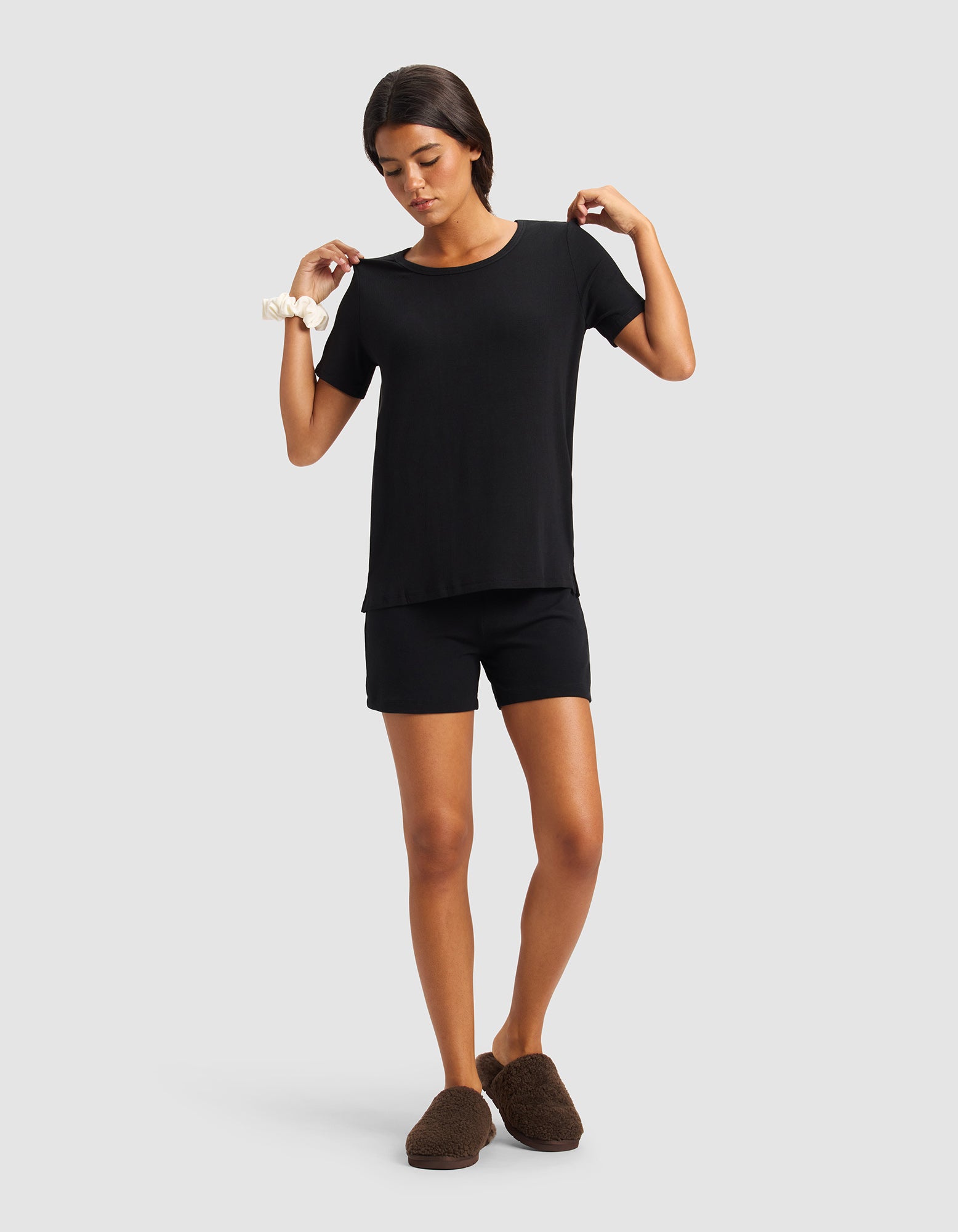 A woman wears the Cozy Earth Women’s Bamboo Rib-Knit Short Sleeve Lounge Top in black, paired with black shorts, brown slippers, and a white scrunchie, standing against a light gray background with her arms raised.