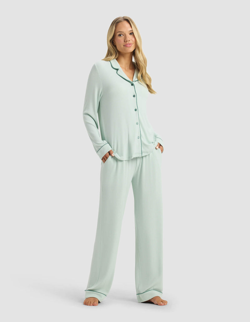 A woman poses barefoot before a plain light background, smiling softly in Cozy Earth’s Women’s Bamboo Rib-Knit Classic Long Sleeve Pajama Set—light blue with button-up top, wide-leg pants, and her hands in the pockets.