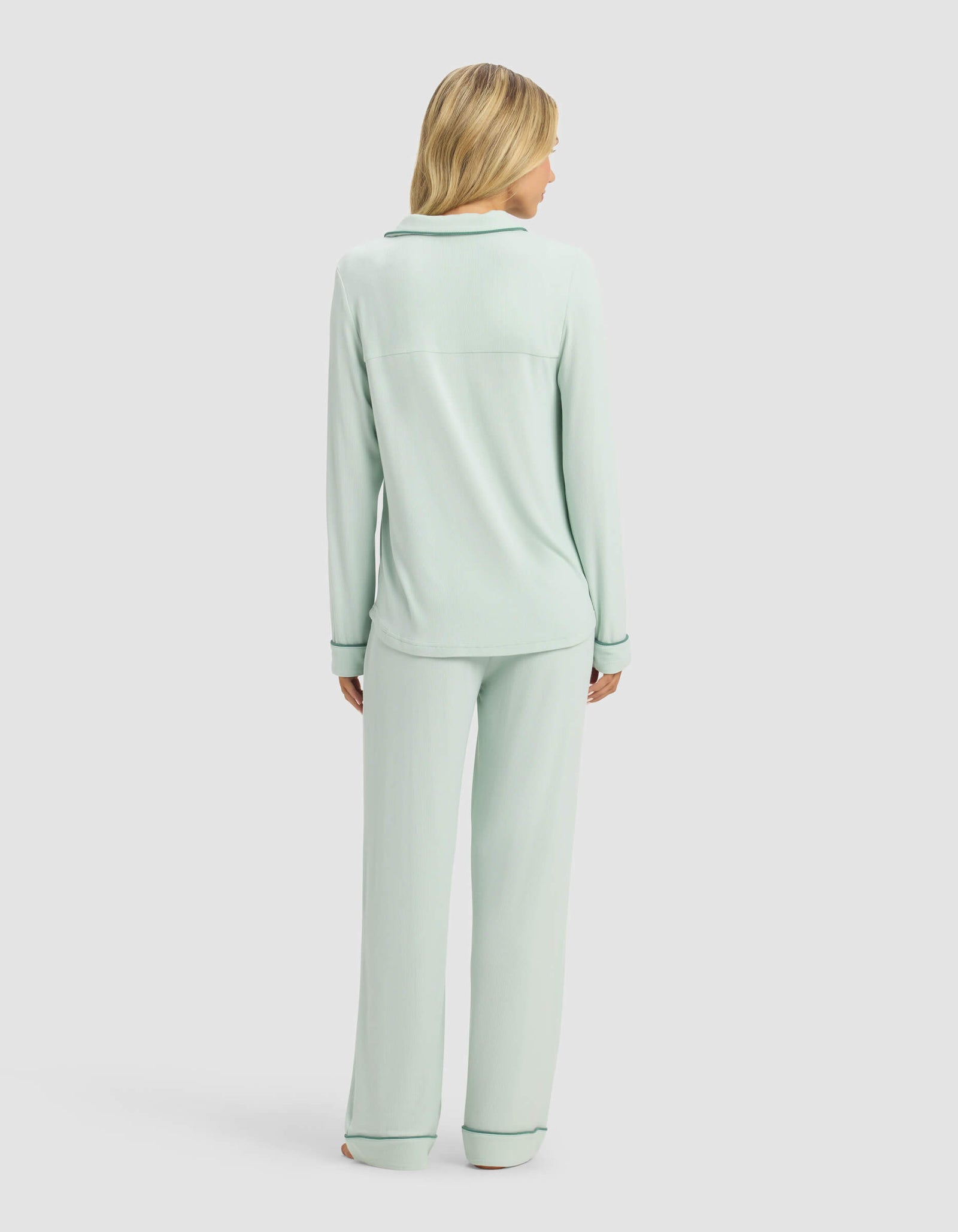 A woman with blonde hair wears the Cozy Earth Women's Bamboo Rib-Knit Classic Long Sleeve Pajama Set in light green, featuring dark piping along the cuffs and collar, as she stands facing away against a plain light background.