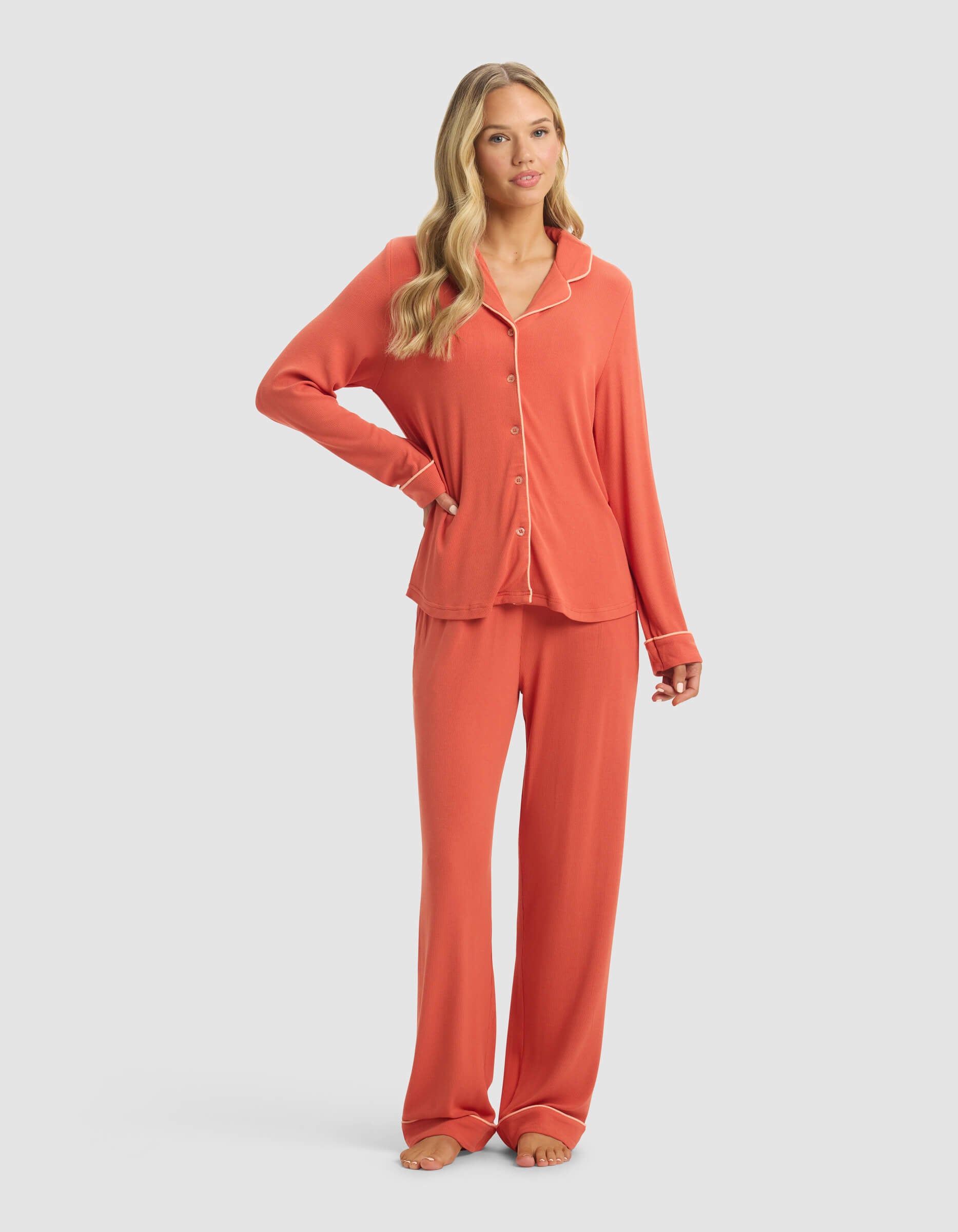 A woman wears the Cozy Earth Women's Bamboo Rib-Knit Classic Long Sleeve Pajama Set, featuring a matching button-up top and long pants in coral. Barefoot, she poses with one hand on her hip against a plain light background.|Color:Sunset