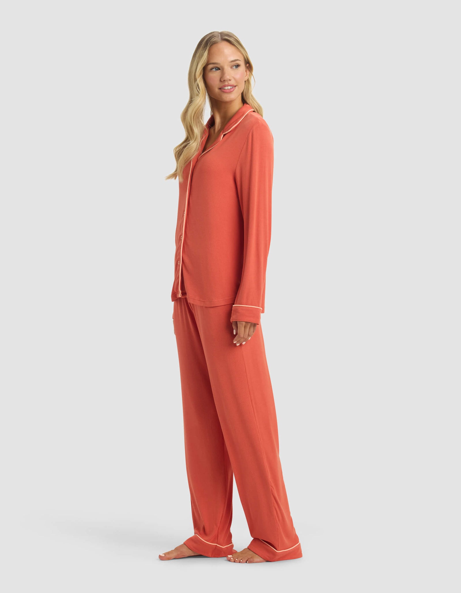 A woman with long, wavy blonde hair smiles gently as she stands barefoot, angled to the side, wearing Cozy Earth's Women's Bamboo Rib-Knit Classic Long Sleeve Pajama Set in coral with white piping, featuring a button-down top and loose pants.