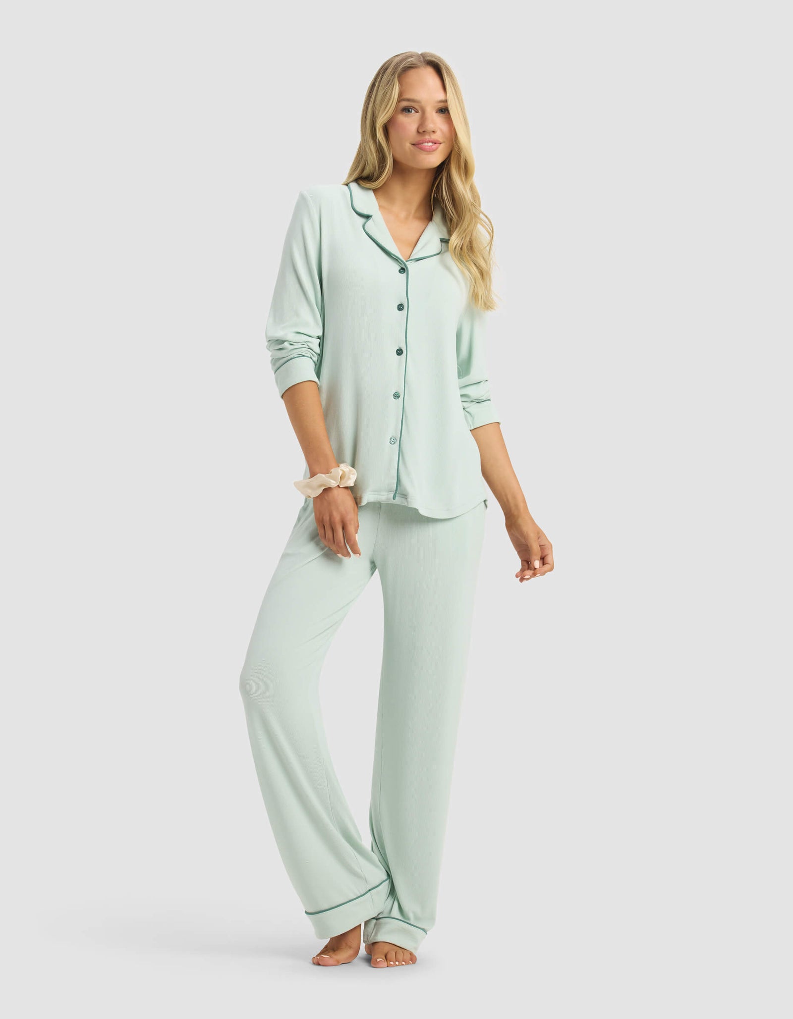 A woman with long blonde hair smiles slightly against a light gray background, wearing a Cozy Earth Women's Bamboo Rib-Knit Classic Long Sleeve Pajama Top in light green and matching long pants, barefoot with a scrunchie on her wrist.