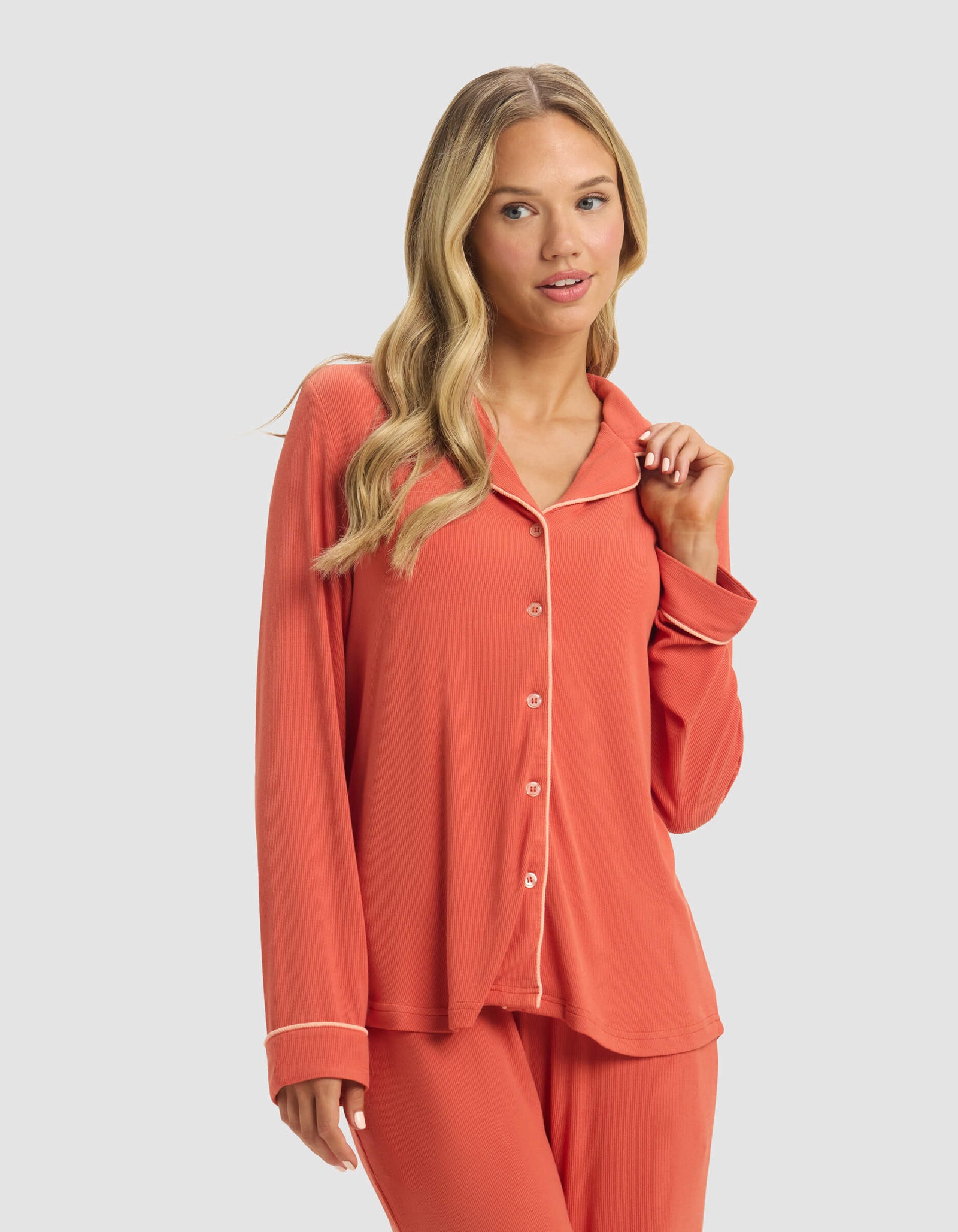 A woman with long blonde hair wears the Cozy Earth Women's Bamboo Rib-Knit Classic Long Sleeve Pajama Top in coral with white piping, paired with matching pants, standing against a plain light gray background.