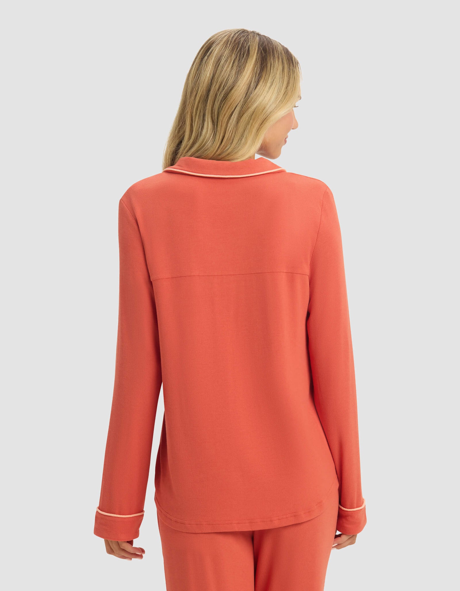 A woman with blonde hair is seen from behind, wearing a coral Women's Bamboo Rib-Knit Classic Long Sleeve Pajama Top by Cozy Earth, standing against a plain light gray background.