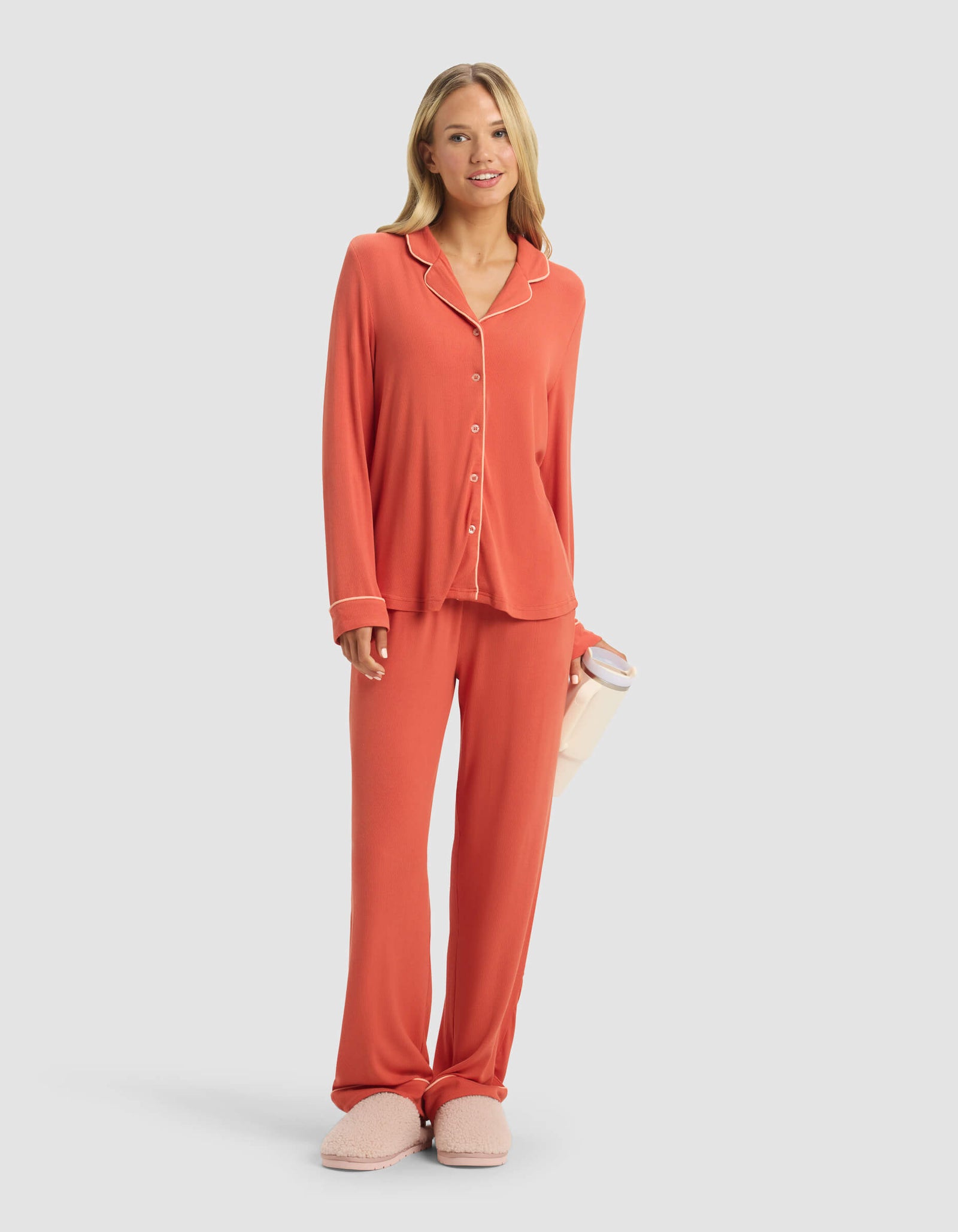 A woman smiles in a Cozy Earth Women's Bamboo Rib-Knit Classic Long Sleeve Pajama Top, paired with matching coral pajama pants, pink slippers, and holding a light pink tumbler. The background is plain light gray.