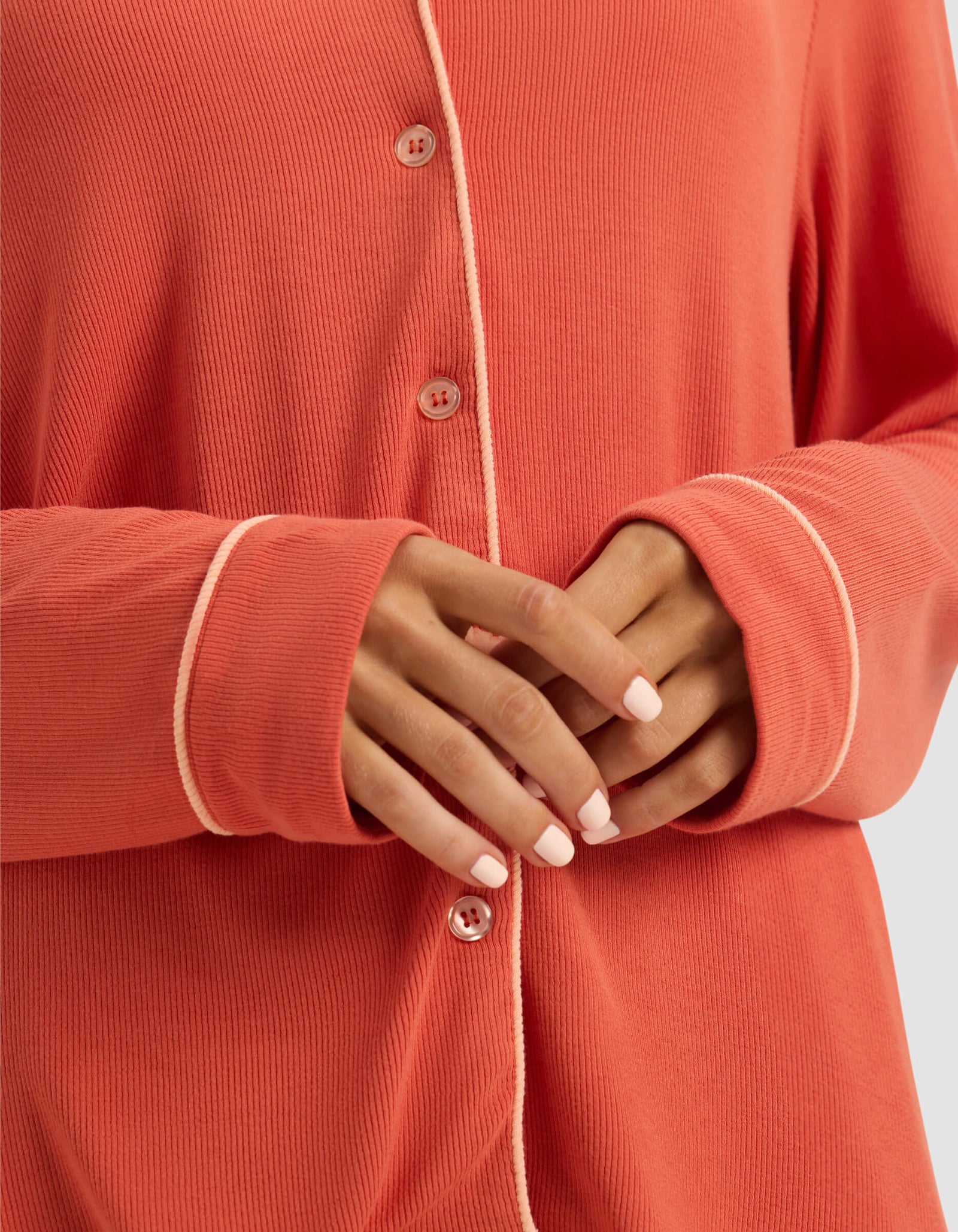 A person with neatly manicured nails softly clasps their hands at their waist while wearing Cozy Earth's Women's Bamboo Rib-Knit Classic Long Sleeve Pajama Top in coral with light trim.