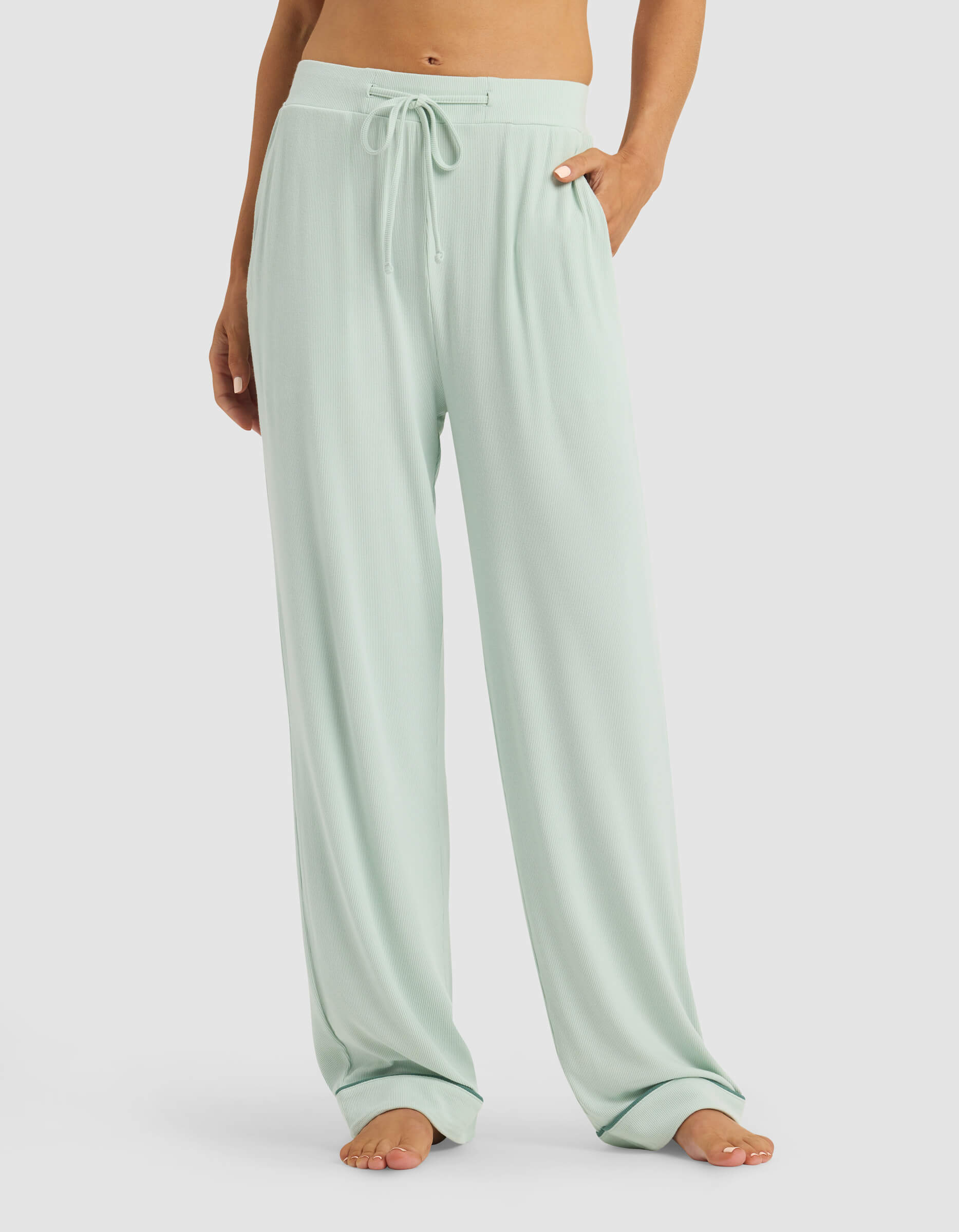 A person stands barefoot with hands in the pockets, wearing Cozy Earth Women's Bamboo Rib-Knit Classic Pajama Pant in light green. Their upper body is cropped out of the image.|Color:Seafoam