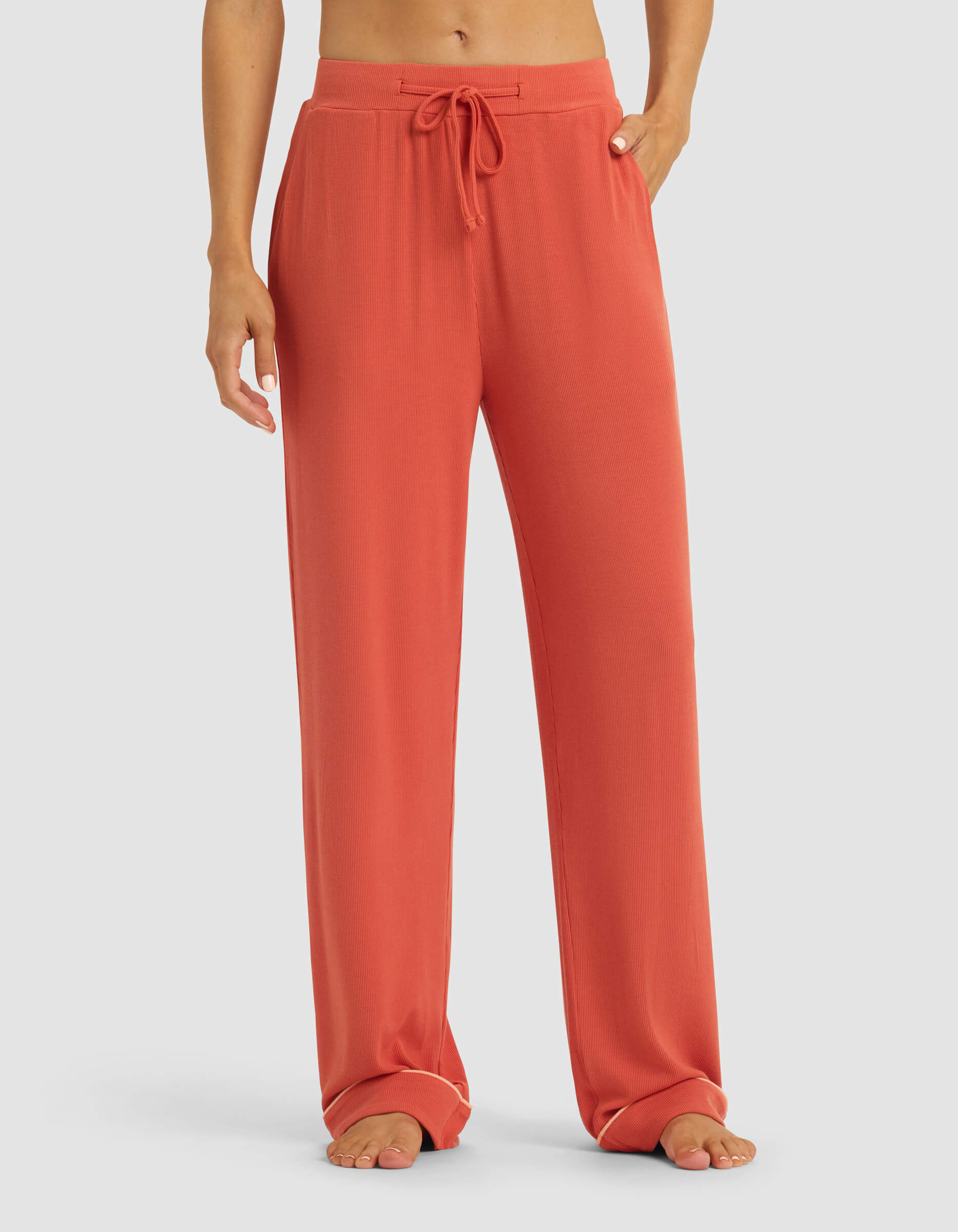 A person stands barefoot against a plain light background wearing Cozy Earth's Women's Bamboo Rib-Knit Classic Pajama Pant in a loose, coral-colored style with a drawstring waist.|Color:Sunset