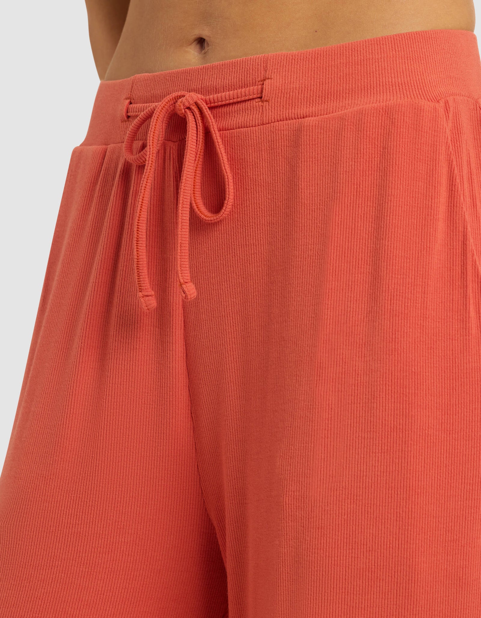 Close-up of a person wearing Cozy Earth Women's Bamboo Rib-Knit Classic Pajama Pant in coral, featuring a high waist, ribbed texture, and drawstring tie. Only the lower torso and upper legs are shown; fabric looks soft and comfortable.
