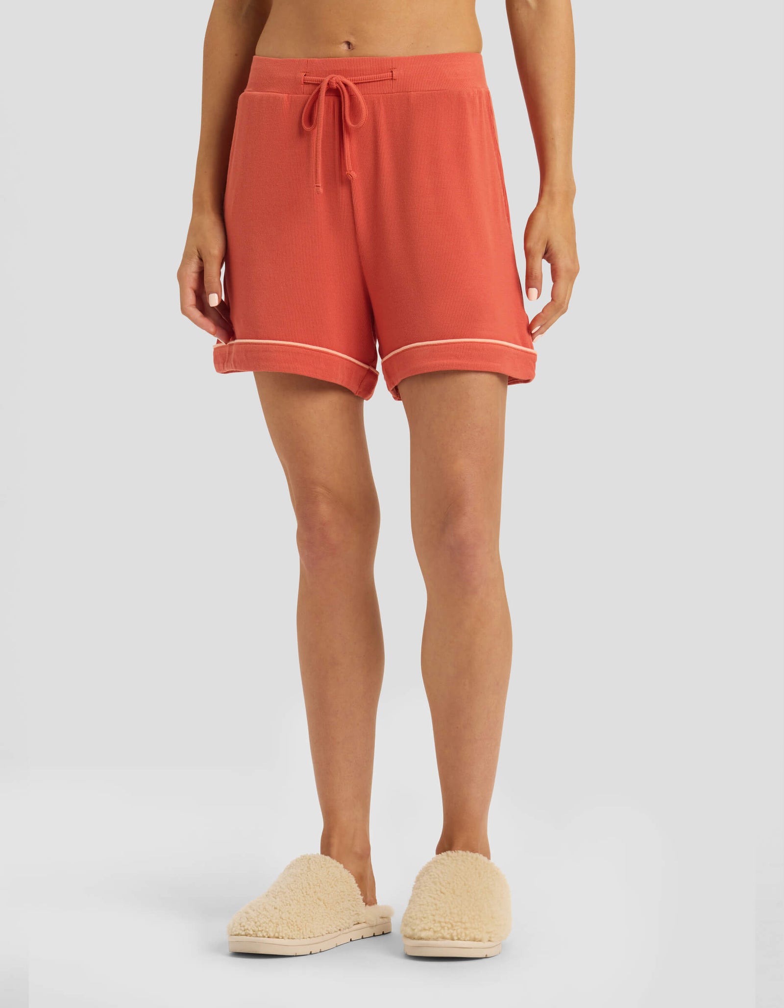 A person is seen from the waist down wearing Cozy Earth Women's Bamboo Rib-Knit Classic Pajama Shorts in coral, featuring a drawstring and rolled hems, paired with cream fuzzy slippers against a plain light gray background.