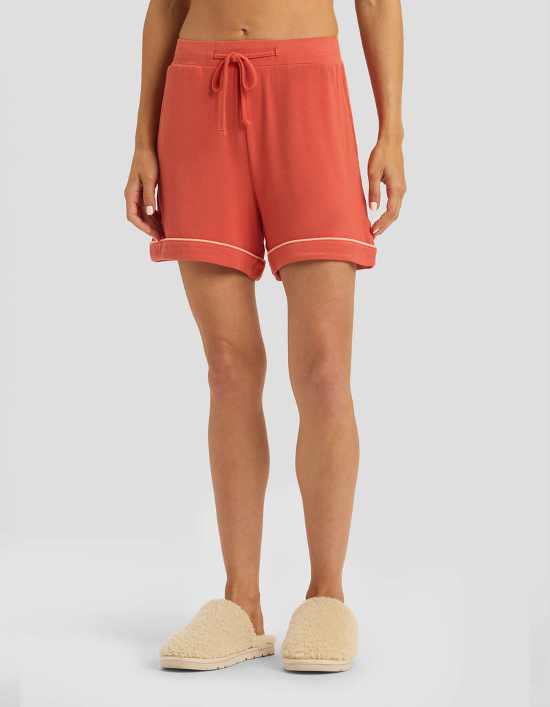 A person is seen from the waist down wearing Cozy Earth Women's Bamboo Rib-Knit Classic Pajama Shorts in coral, featuring a drawstring and rolled hems, paired with cream fuzzy slippers against a plain light gray background.