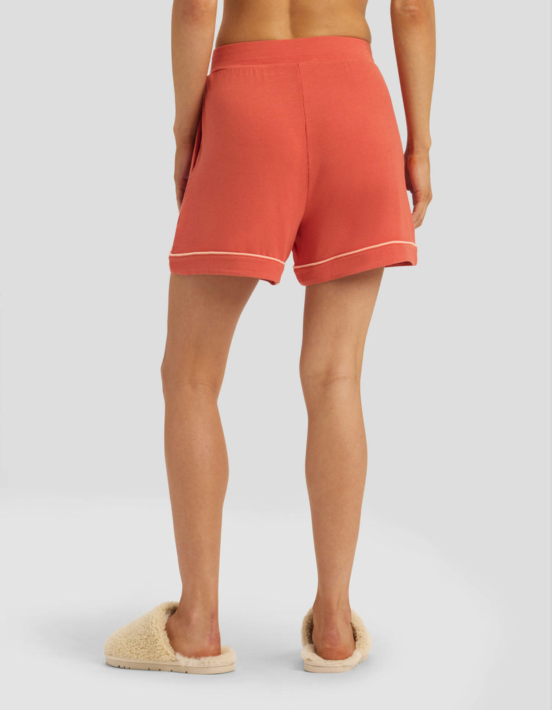 A person stands facing away on a plain light background, wearing Cozy Earth Women's Bamboo Rib-Knit Classic Pajama Shorts in coral with white trim and cream fuzzy slippers.