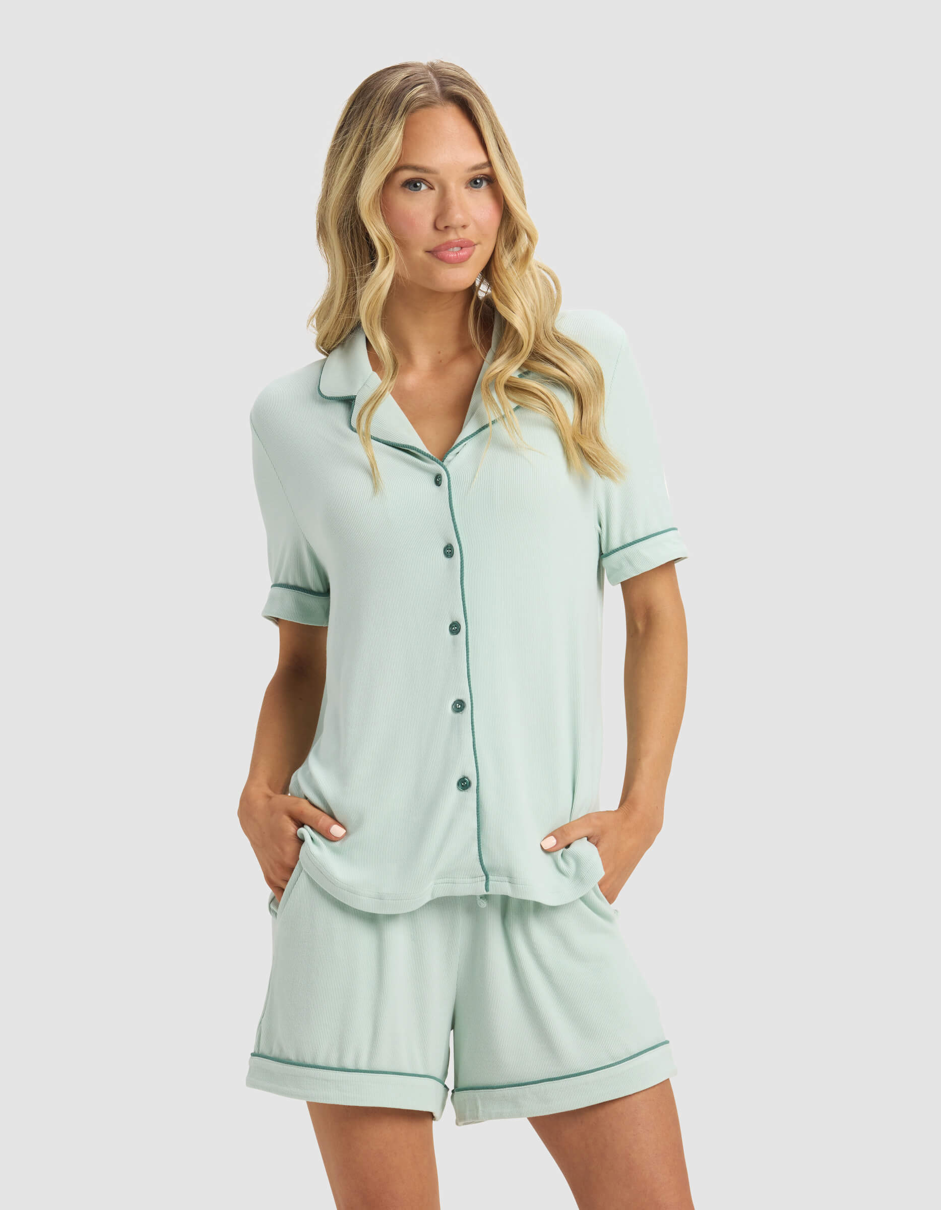 A woman with long, wavy blonde hair wears the Cozy Earth Women's Bamboo Rib-Knit Classic Short Sleeve Pajama Top in light green, paired with matching shorts. She stands against a plain light background with her hands in her pockets.|Color:Seafoam
