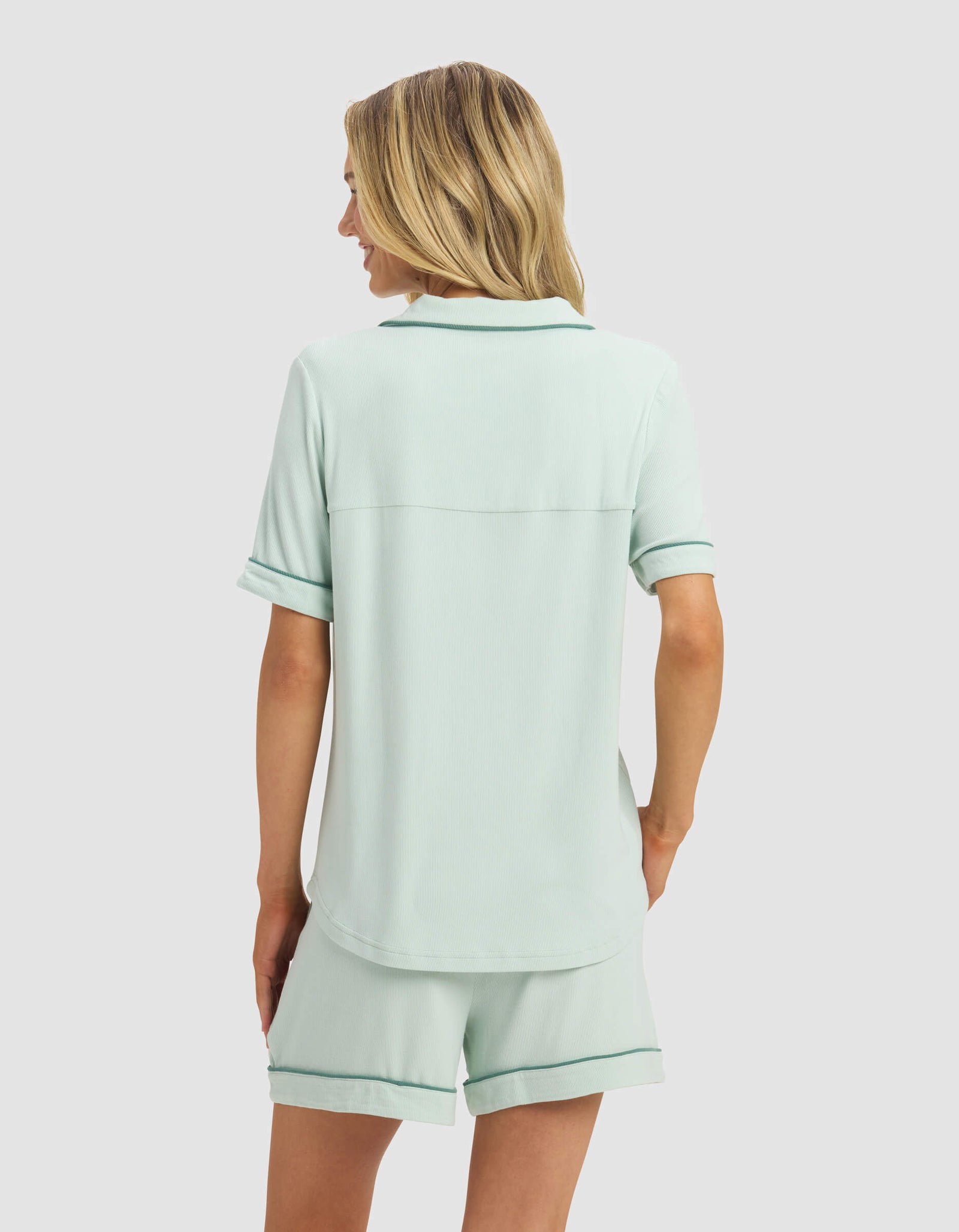 A woman with blonde hair is seen from the back, wearing Cozy Earth's Women's Bamboo Rib-Knit Classic Short Sleeve Pajama Top in light green and matching relaxed-fit shorts, standing against a plain light gray background.