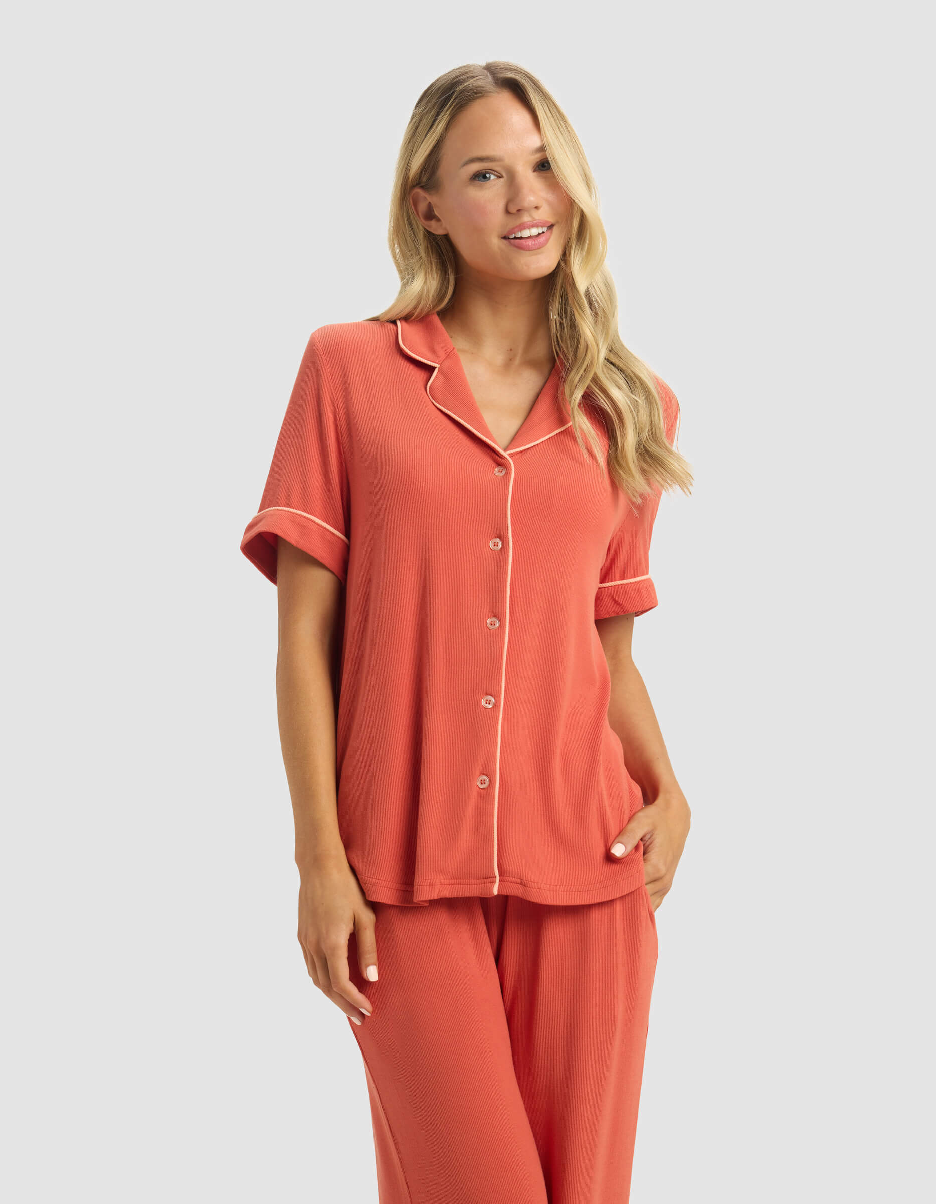A woman with long blonde hair smiles with one hand in her pocket, wearing the Cozy Earth Women's Bamboo Rib-Knit Classic Short Sleeve Pajama Top and matching pants against a light gray background.|Color:Sunset
