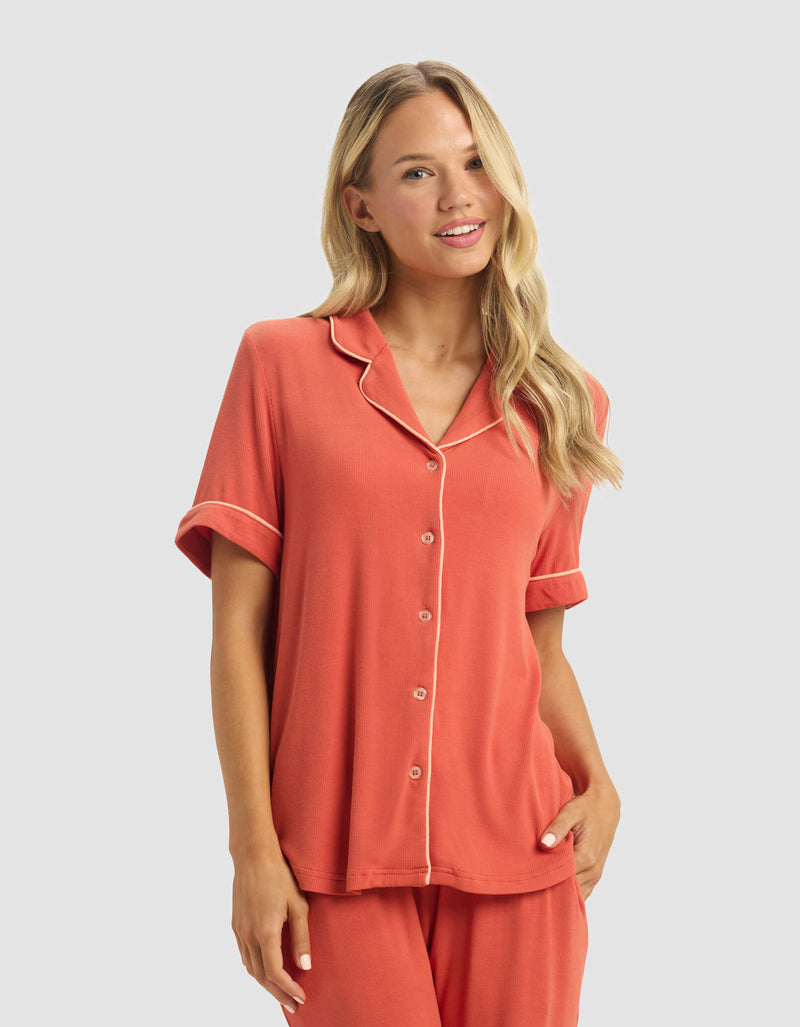 A woman with long blonde hair smiles in the Cozy Earth Women's Bamboo Rib-Knit Classic Short Sleeve Pajama Set, a coral ensemble with white piping, button-up top, and matching pants, standing against a plain light background.