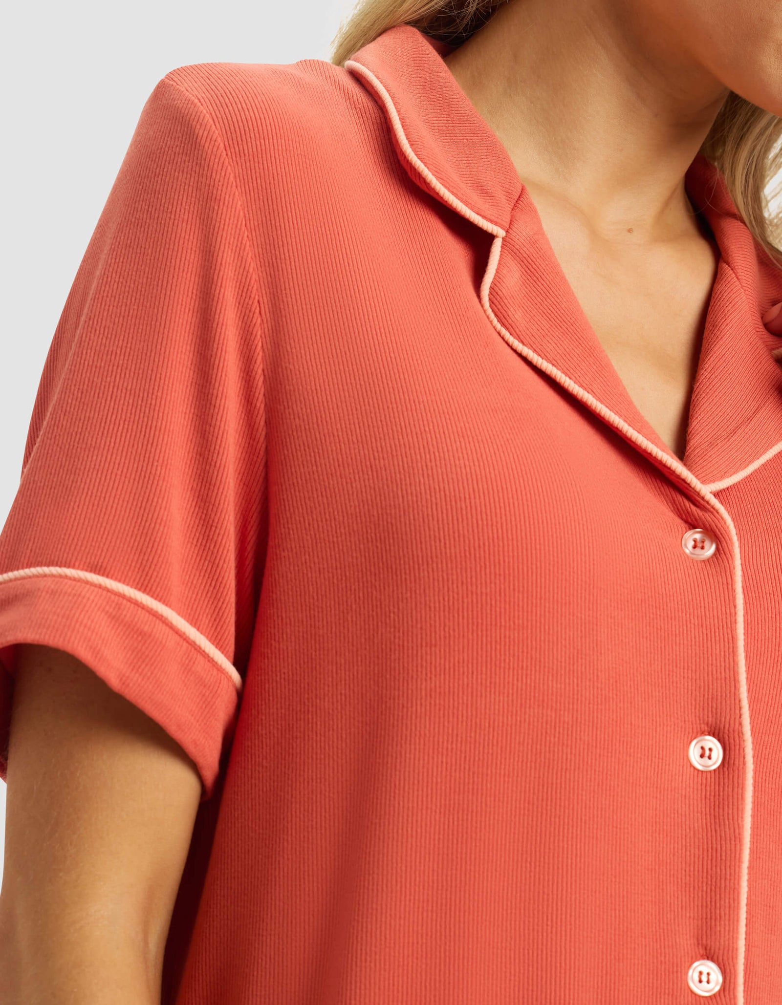 A close-up shows a person wearing the Cozy Earth Women's Bamboo Rib-Knit Classic Short Sleeve Pajama Top in coral, featuring white piping on the collar and sleeves, against a light gray background. The image highlights the upper chest, shoulder, and part of the face.