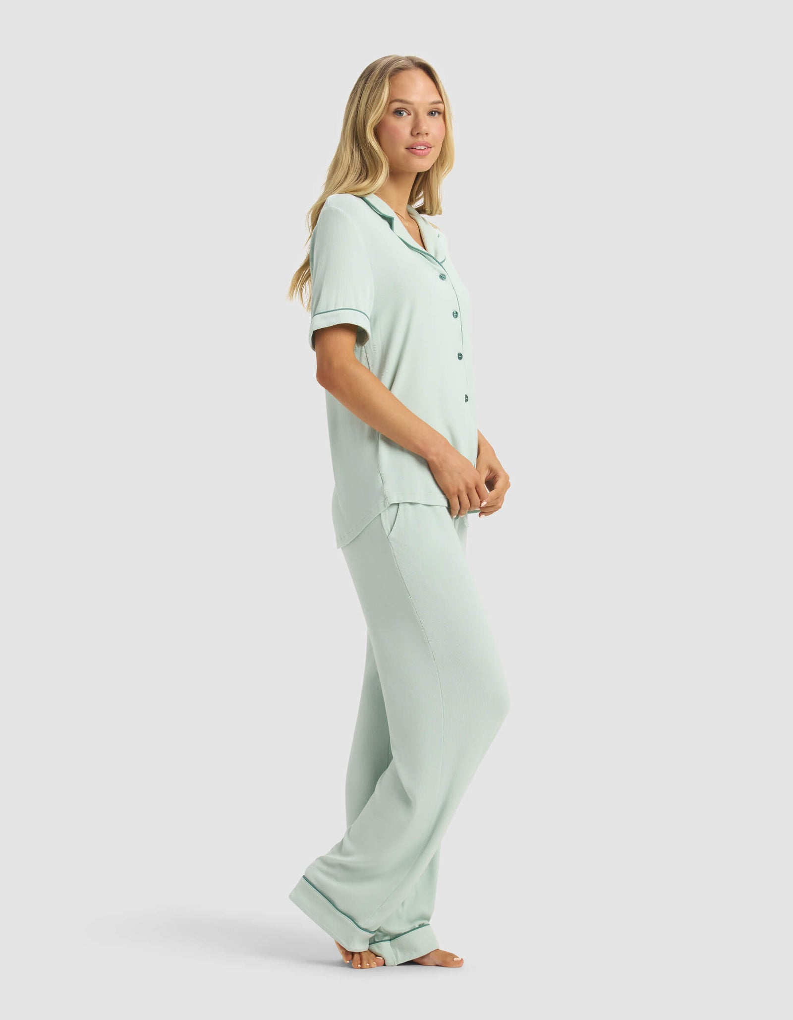 A woman stands barefoot in the Cozy Earth Women's Rib-Knit Classic Short Sleeve & Pant Pajama Set, featuring a light blue button-up top and pants. She poses side-on, looking at the camera against a plain light background.
